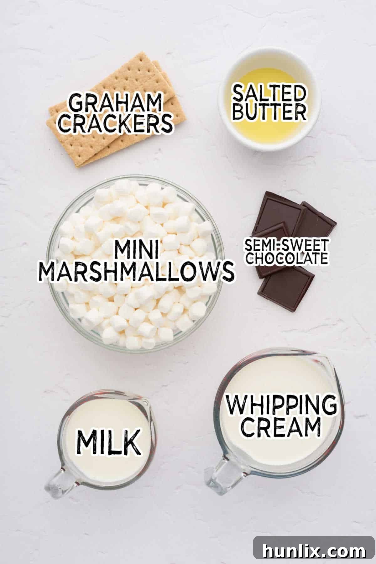 A selection of fresh ingredients including mini marshmallows, milk, whipping cream, chocolate, graham crackers, and butter, artfully arranged on a clean white background, ready for pie preparation.