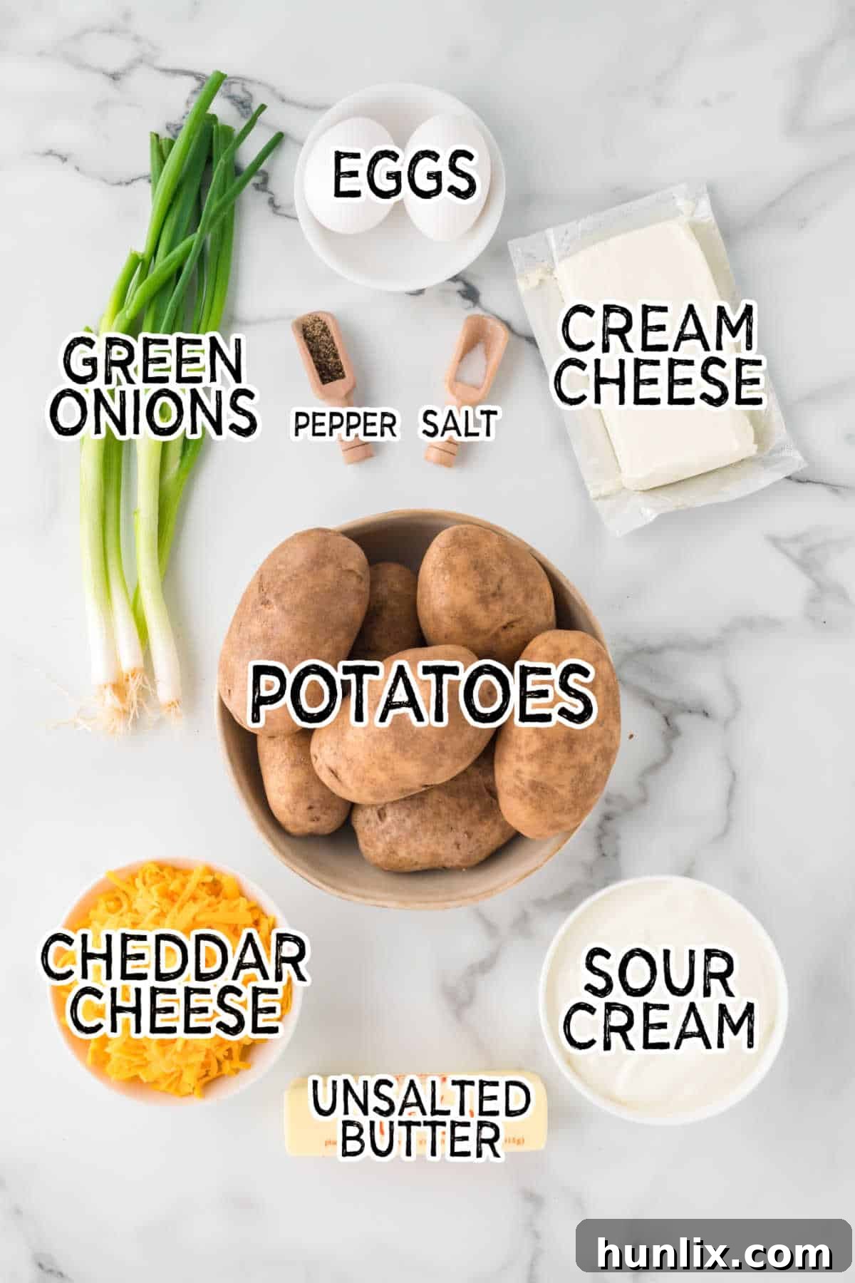 Various ingredients for mashed potato casserole on a white marble counter, including potatoes, cheeses, sour cream, and butter.