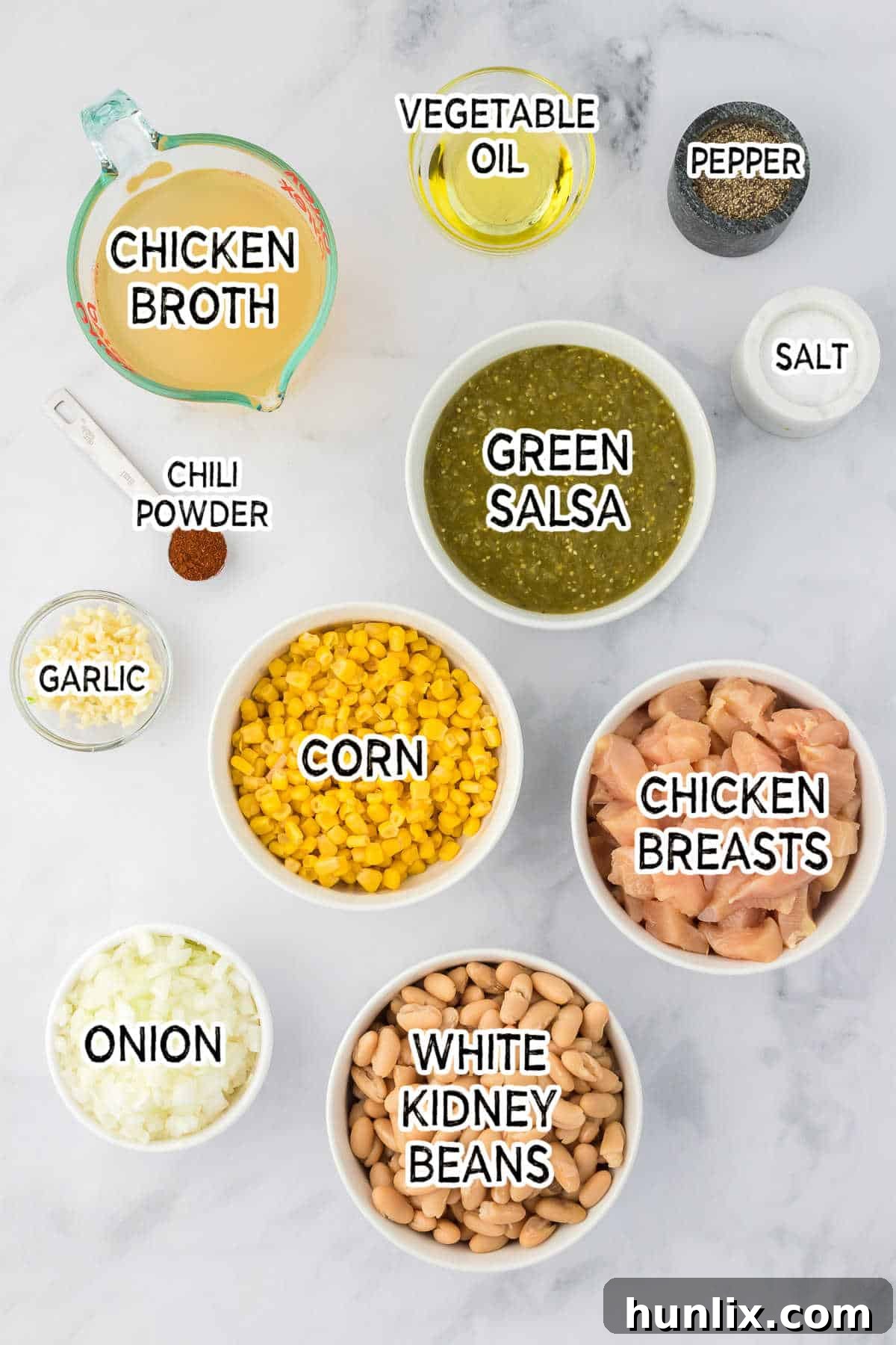 Assorted fresh ingredients neatly arranged on a counter, including chicken breasts, onions, garlic, white kidney beans, green salsa, and corn, ready for making white chicken chili.