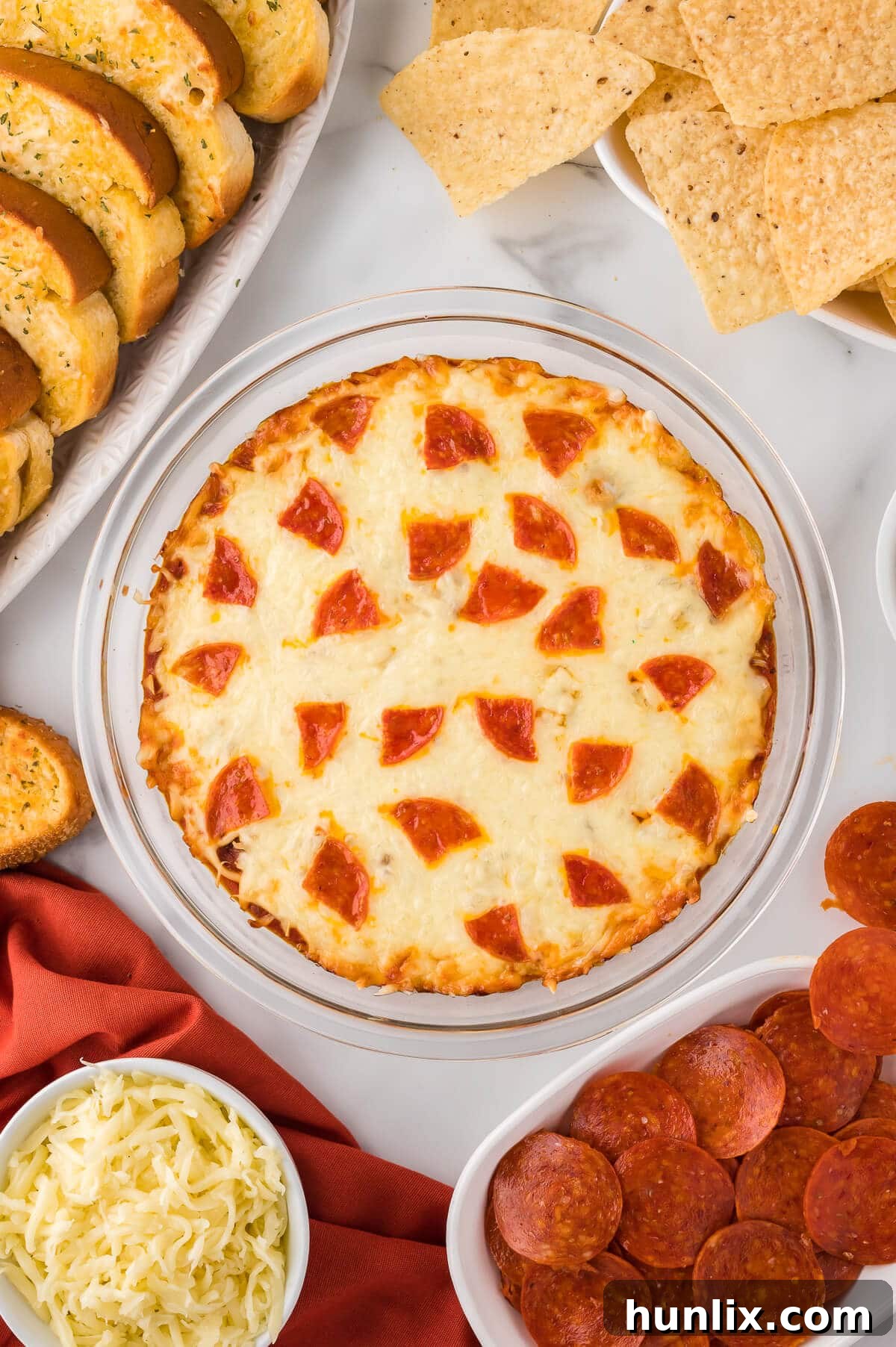 A bubbling hot pizza dip in a rustic pie pan, surrounded by fresh ingredients like pepperoni, olives, and crisp breadsticks, ready to be served at a party.