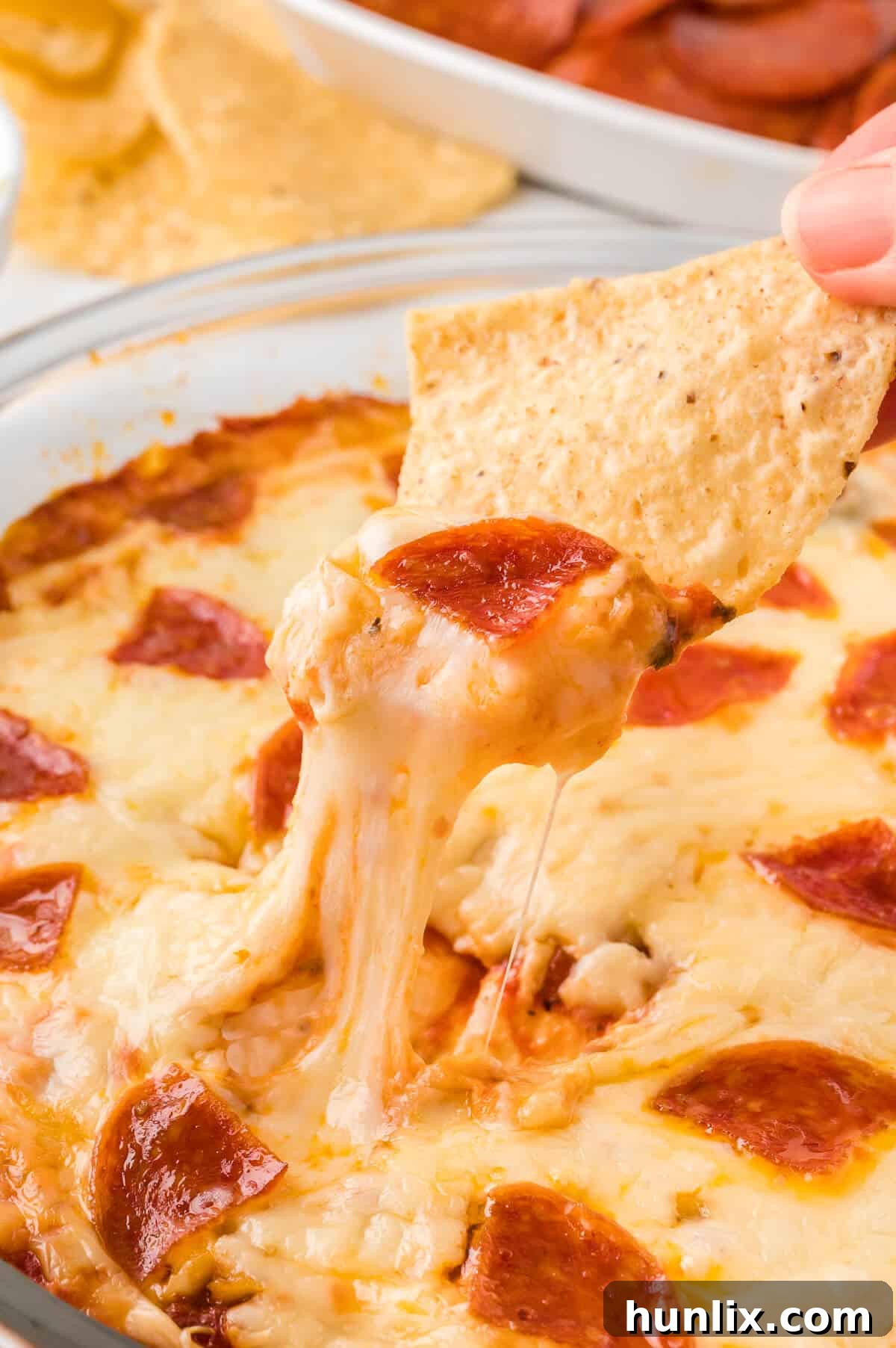A hand enthusiastically dipping a crispy tortilla chip into the warm, gooey, and cheesy pizza dip, creating an irresistible stretch of mozzarella.