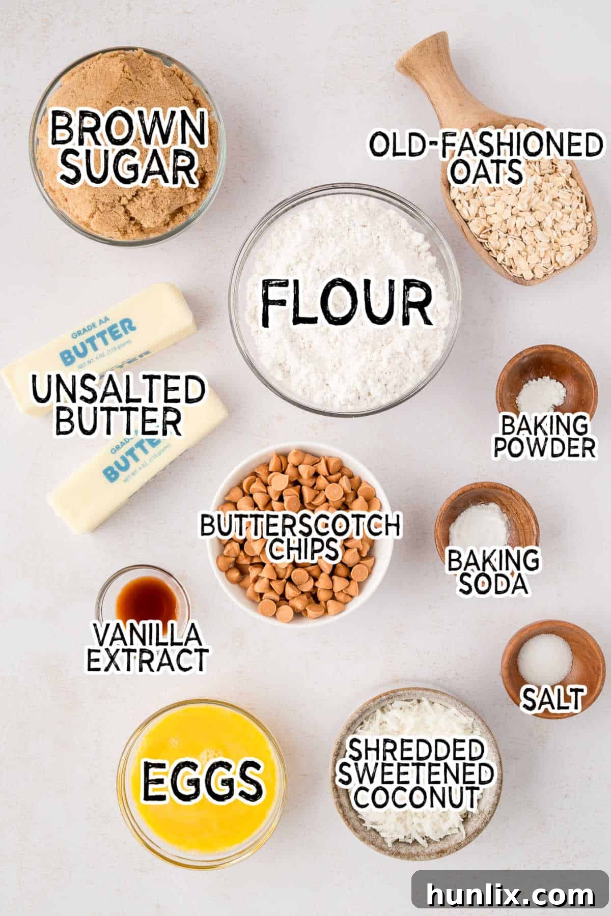 A collection of ingredients for Coconut Butterscotch Cookies laid out, including flour, sugar, butter, eggs, oats, coconut, and butterscotch chips.