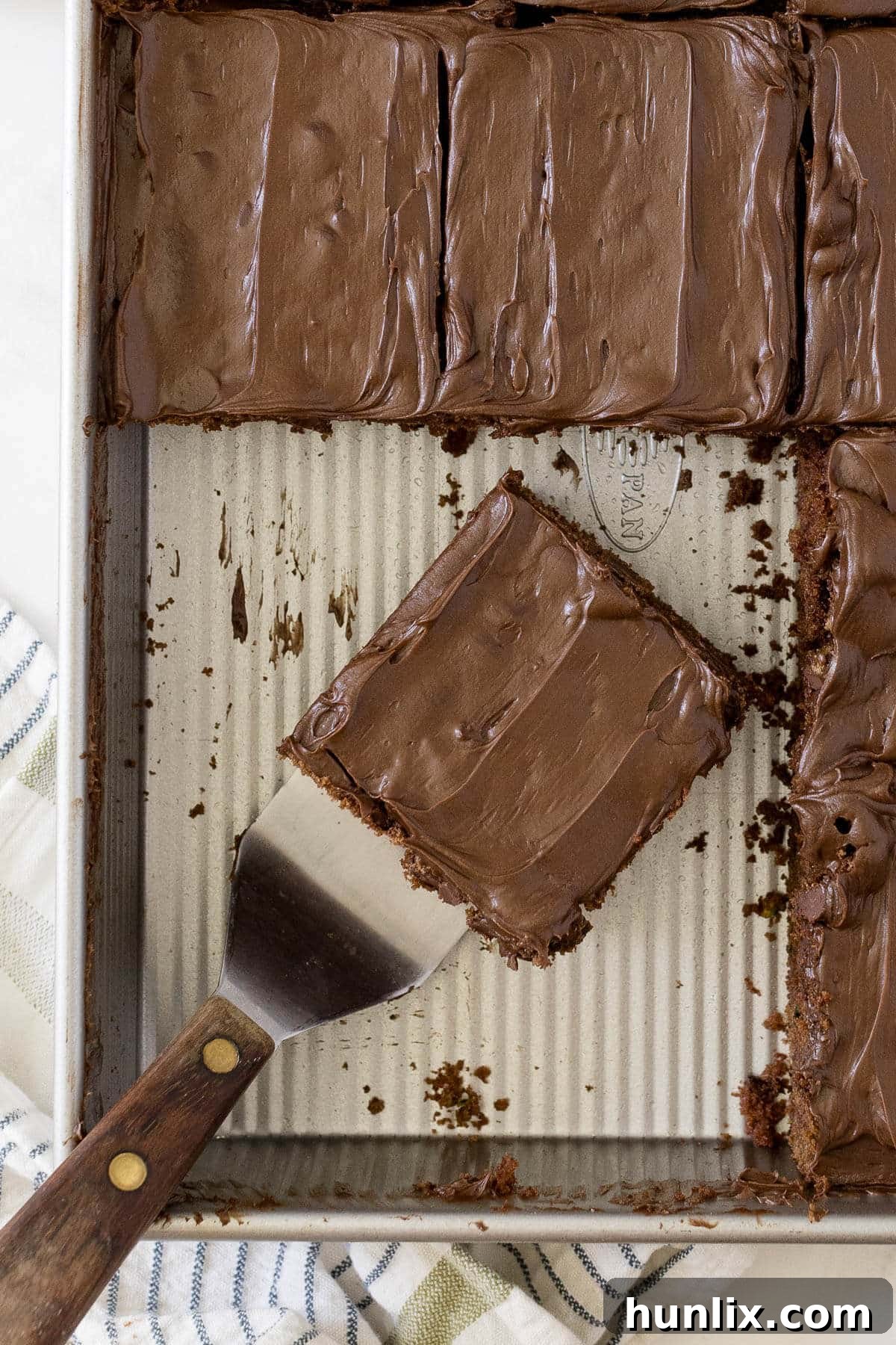 A generous slice of moist chocolate zucchini cake, adorned with melted chocolate chips, resting in a baking pan beside a serving spatula. The cake has a rich, dark brown color, indicating a deep chocolate flavor.