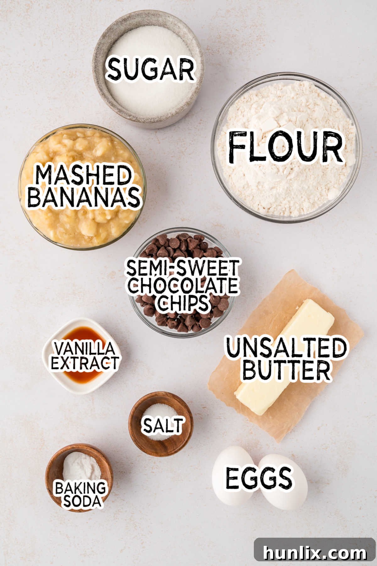 Decadent Banana Chocolate Chip Muffins 3 A collection of baking ingredients including bananas, chocolate chips, butter, eggs, flour, and sugar, neatly arranged on a white surface.