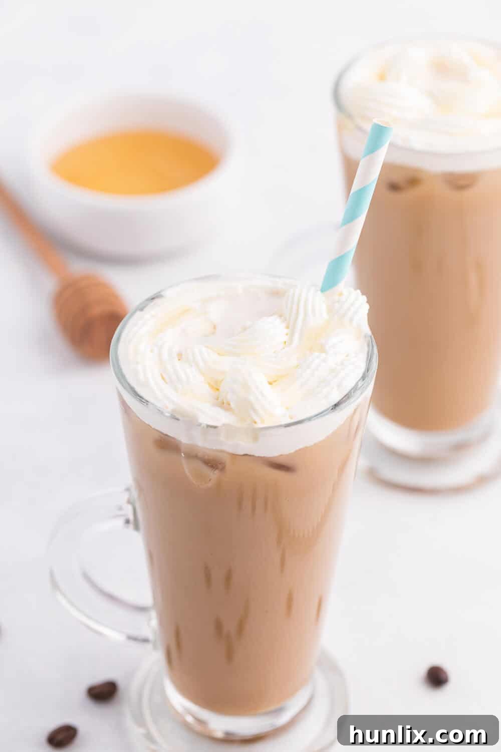 A perfectly crafted, frosty glass of Iced Honey Almond Milk Latte with a golden straw, highlighting its refreshing appeal. The latte is topped with a generous swirl of whipped cream and a delicate drizzle of honey.