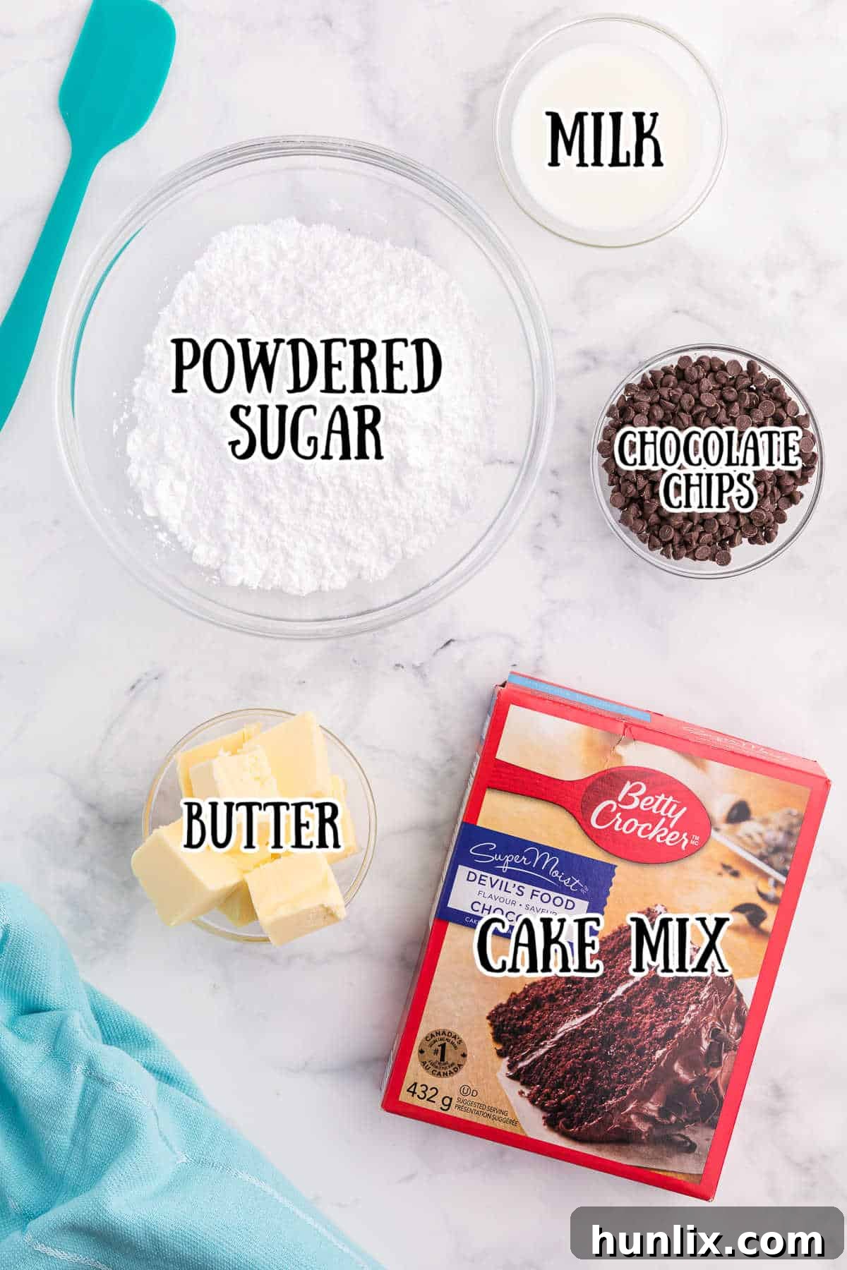 A selection of ingredients for chocolate cake batter fudge, including cake mix, powdered sugar, butter, milk, and chocolate chips, arranged on a white marble background.