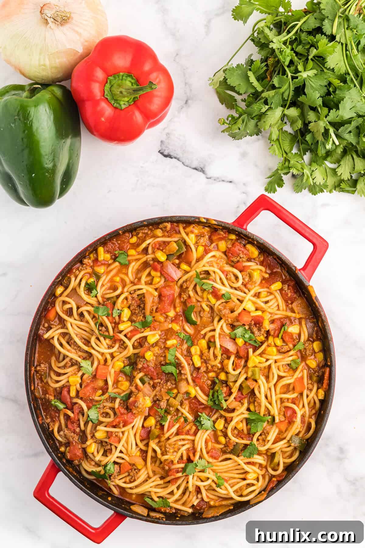 A large pan filled with vibrant Mexican spaghetti, surrounded by fresh ingredients like bell peppers, corn, and tomatoes.