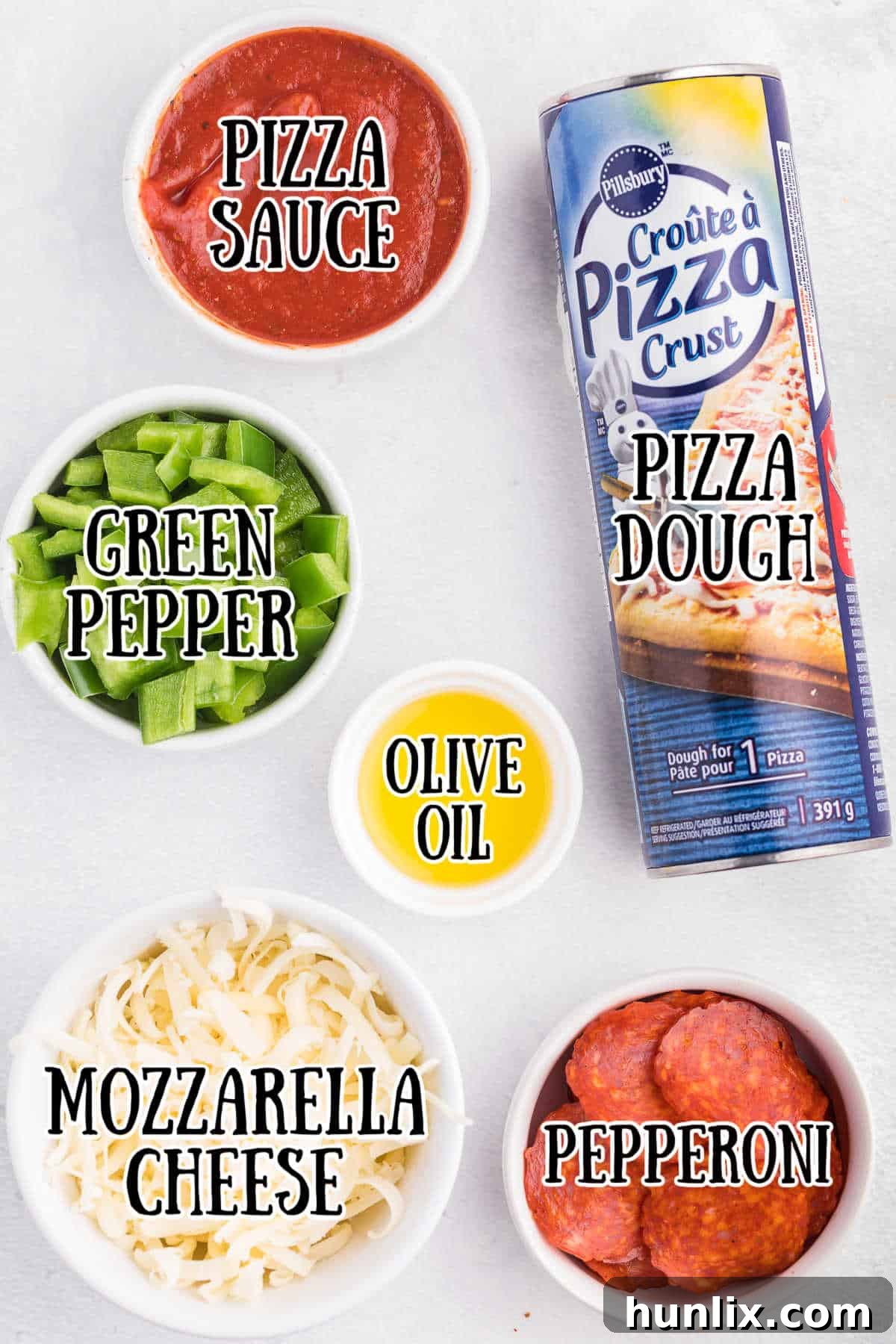 A selection of fresh pizza ingredients laid out on a white background: a can of pizza dough, pepperoni slices, chopped green pepper, a bowl of shredded mozzarella cheese, and a bottle of olive oil.
