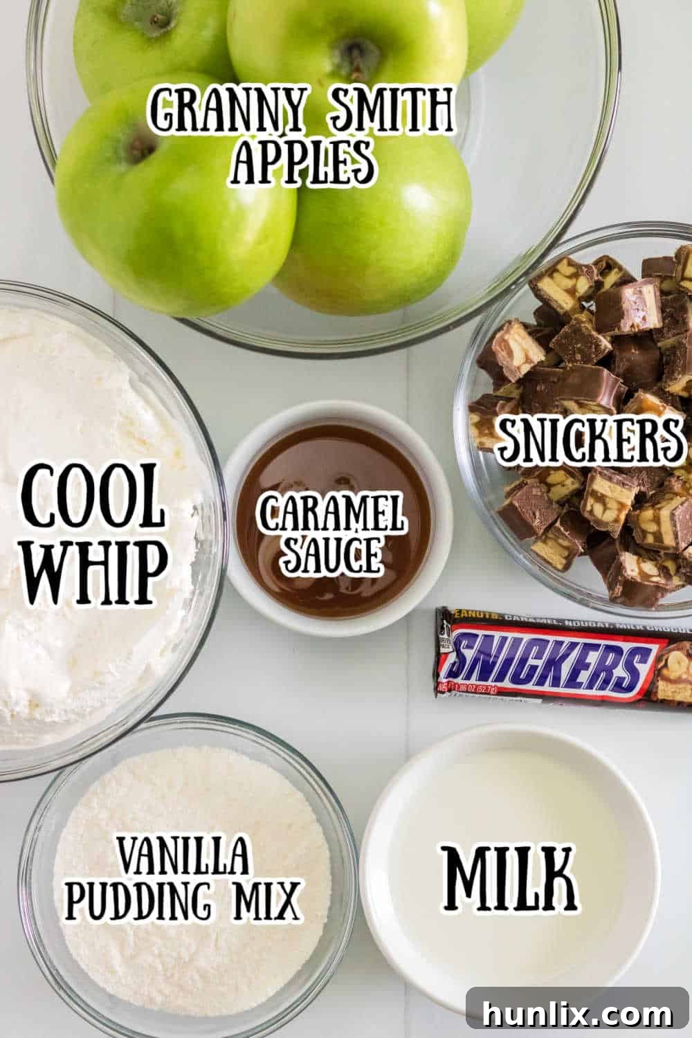Creamy Caramel Snickers Apple Salad 3 All the fresh ingredients for Snickers Salad, including vanilla pudding mix, milk, Cool Whip, Granny Smith apples, Snickers bars, and caramel sauce, neatly arranged on a white wooden surface.