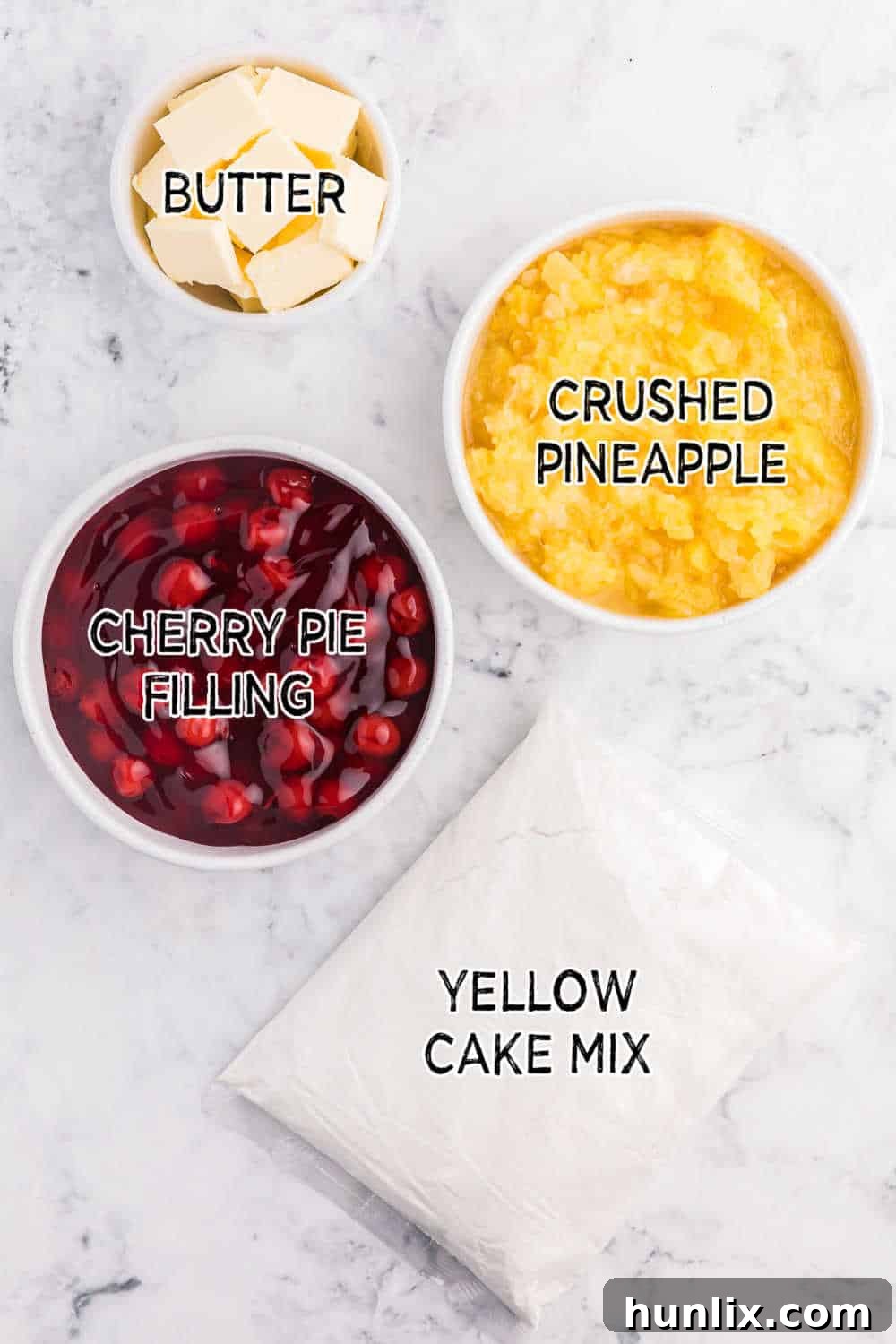 Ingredients for Cherry Pineapple Dump Cake laid out on a white marble background: cans of crushed pineapple and cherry pie filling, a box of yellow cake mix, and a stick of butter.
