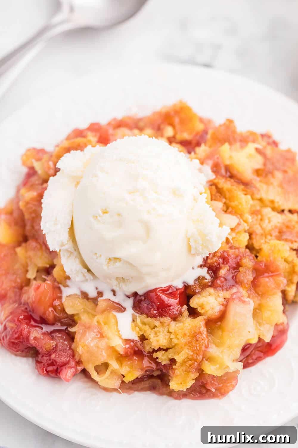 A slice of Cherry Pineapple Dump Cake topped with melting vanilla ice cream on a white plate.