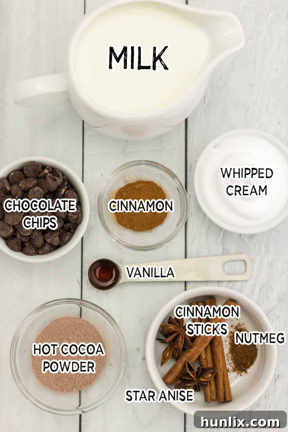 Assortment of Cinnamon Hot Chocolate ingredients laid out on a white wooden background, including milk, chocolate chips, cocoa powder, cinnamon sticks, vanilla extract, and star anise.