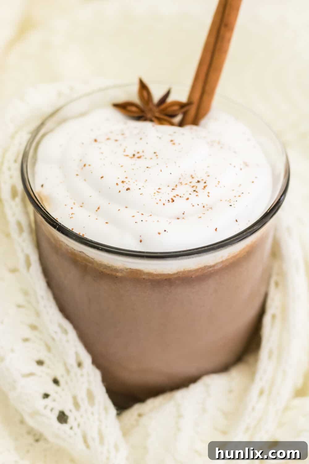 Cinnamon hot chocolate served in a clear glass mug, nestled in a cream knitted blanket, creating a cozy winter scene.