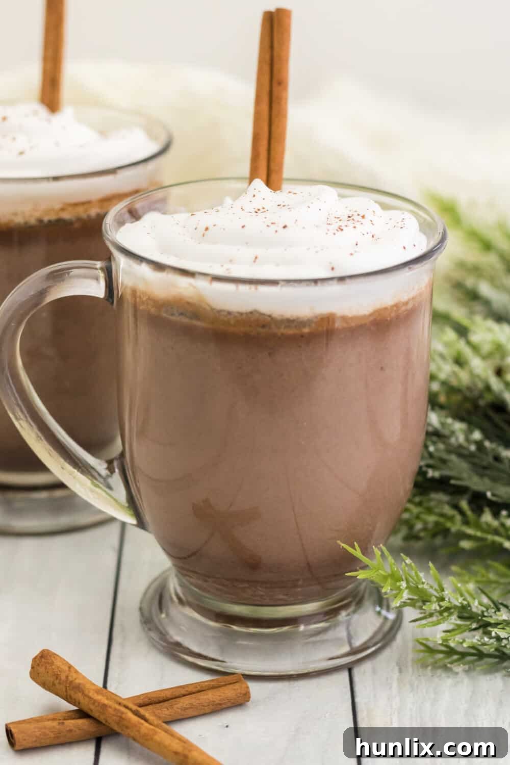 Another view of cinnamon hot chocolate in a glass mug, with whipped cream and a cinnamon stick garnish, emphasizing its inviting texture.