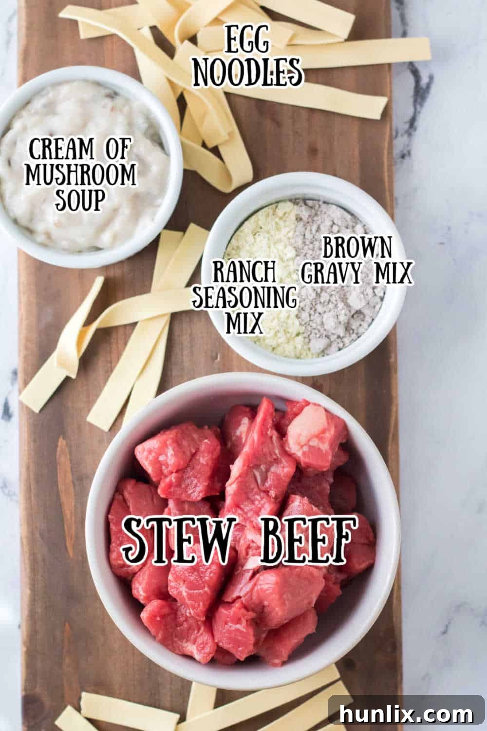 A flat lay of all the ingredients needed to make slow cooker beef and noodles, including stew beef, seasoning packets, cream of mushroom soup, and egg noodles.