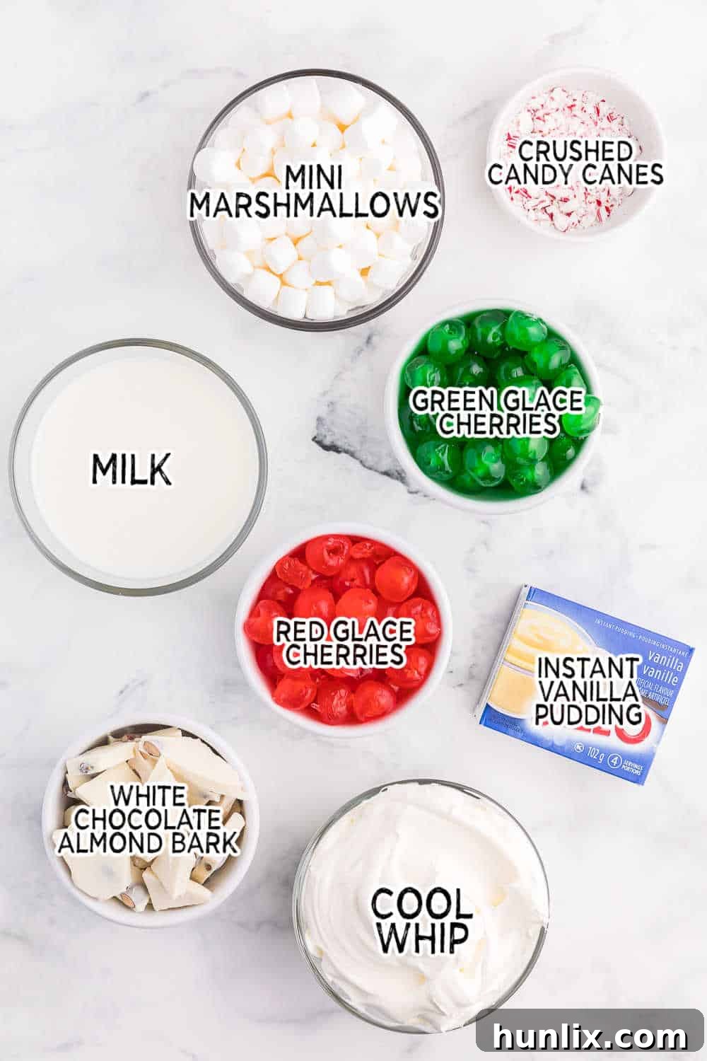 A collection of ingredients laid out on a white surface, including instant pudding mix, milk, marshmallows, glace cherries, white chocolate almond bark, Cool Whip, and candy canes, ready to make Christmas Fluff.