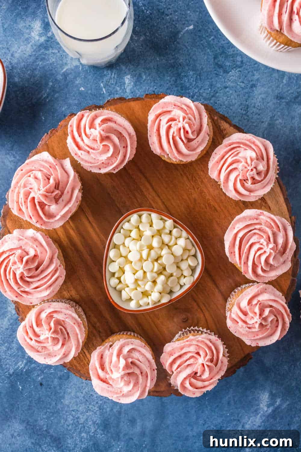 Sweet strawberry cupcakes topped with pink strawberry buttercream frosting and white chocolate chips, arranged on a circular wooden platter next to a small bowl of extra white chocolate chips.