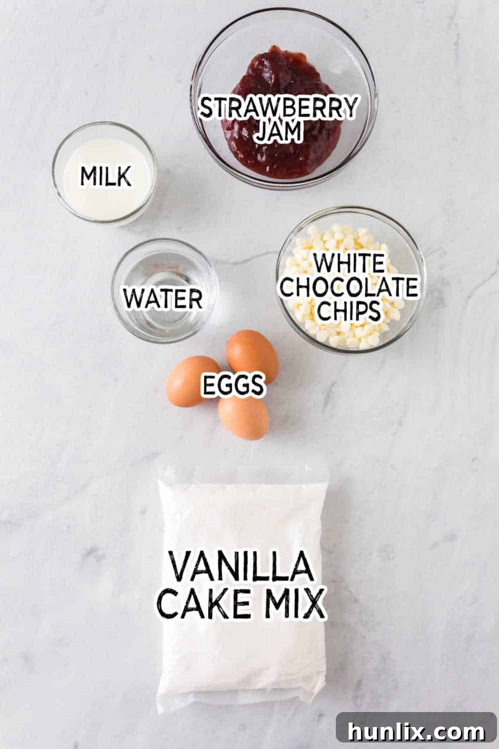 Various ingredients for strawberry cupcakes laid out on a white marble background, including a box of cake mix, eggs, milk, water, strawberry jam, and white chocolate chips.