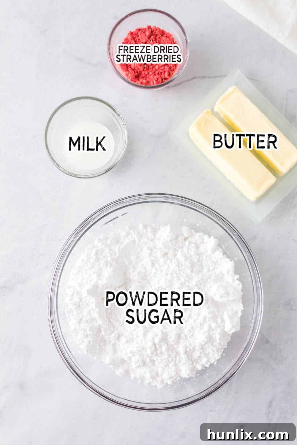 Ingredients for strawberry buttercream frosting, including powdered sugar, freeze-dried strawberries, softened butter, milk, and vanilla extract, arranged on a white background.