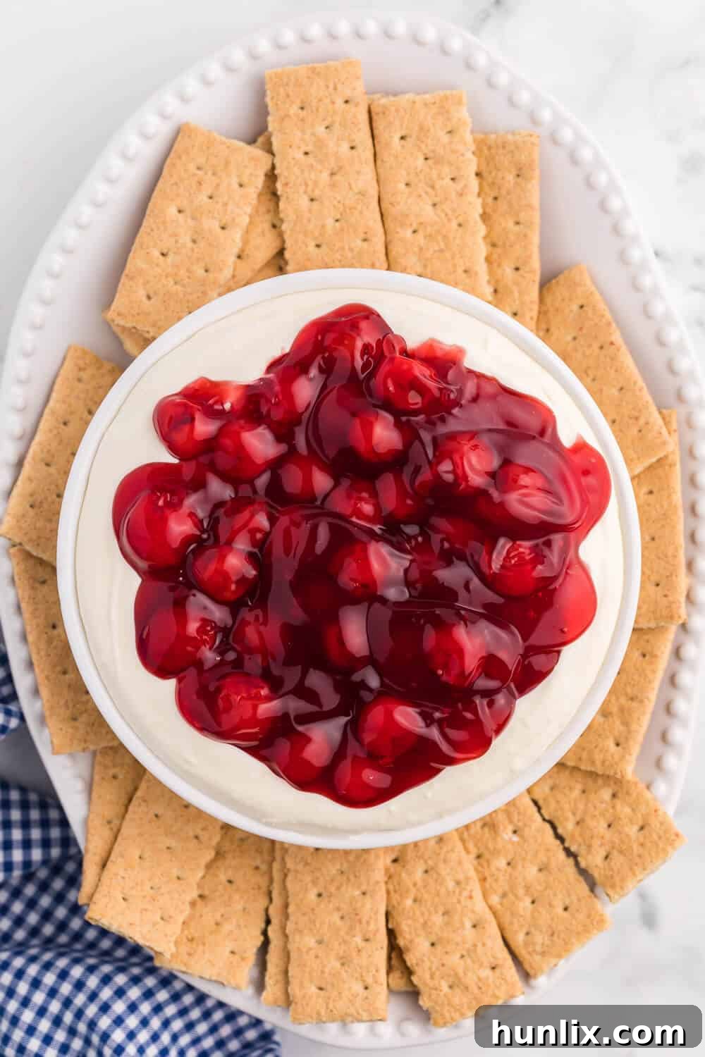 Vibrant cherry cheesecake dip on a white platter, garnished with fresh mint and surrounded by an assortment of graham crackers, vanilla wafers, and shortbread cookies, inviting a delicious tasting experience.