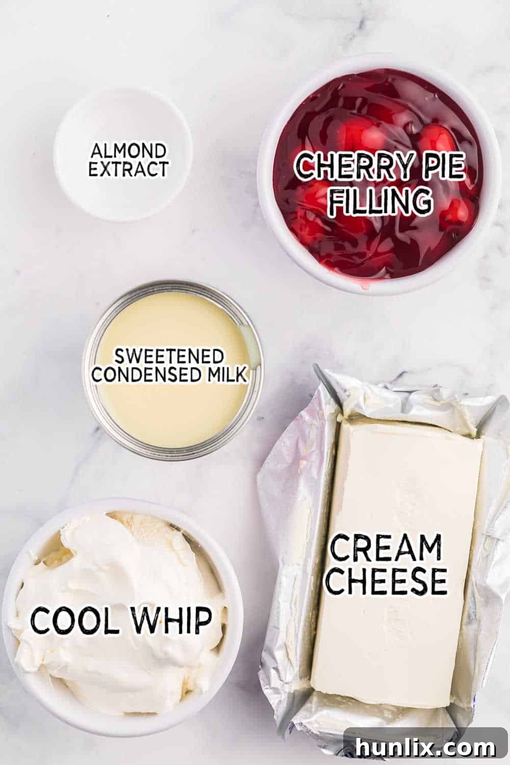 A collection of ingredients neatly arranged on a counter, including a block of cream cheese, a can of sweetened condensed milk, a tub of whipped topping, a bottle of almond extract, and a can of cherry pie filling, ready for making a delicious dip.