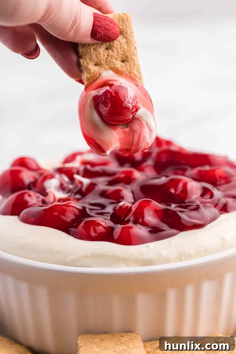 A hand dipping a graham cracker into a creamy cherry cheesecake dip, highlighting the perfect texture and the rich cherry topping, ready for a delicious bite.