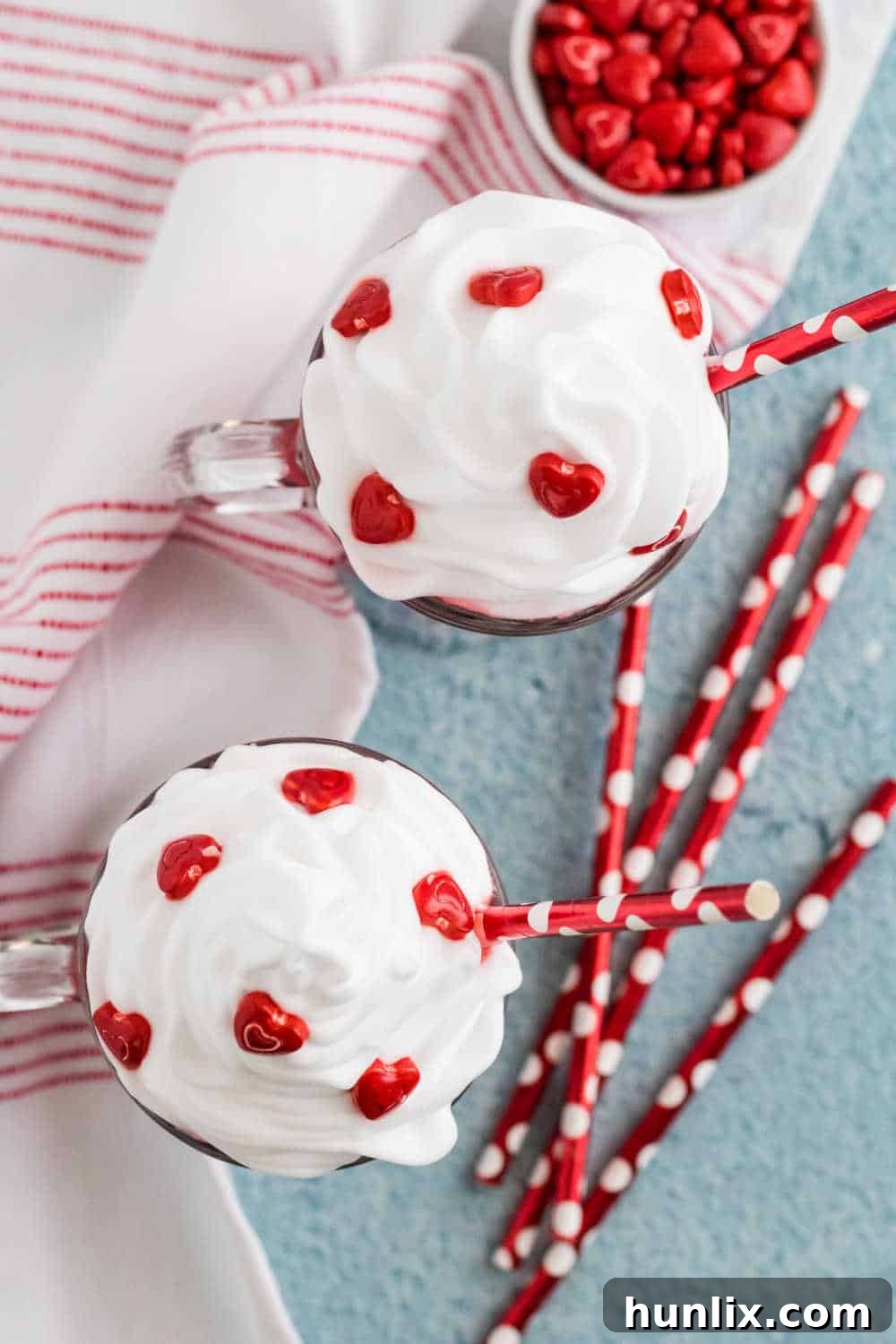 Two mugs of red velvet hot chocolate, each with a red striped straw, topped with whipped cream and heart sprinkles, set on a rustic surface.