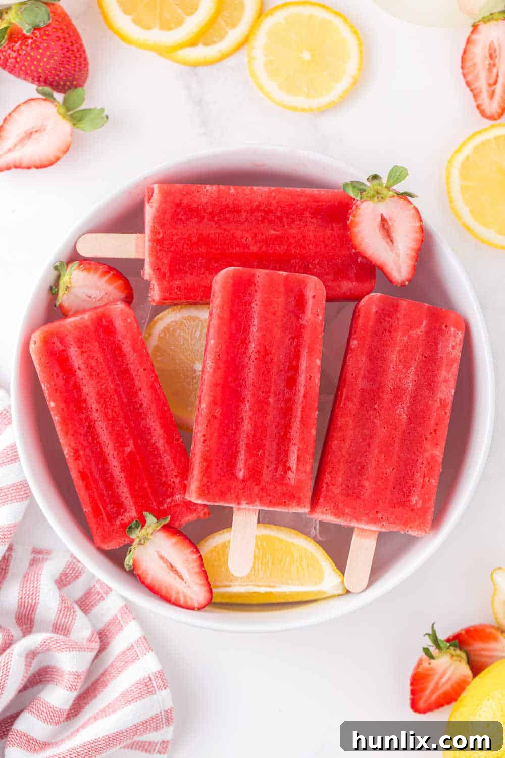 A bowl of ice filled with vibrant pink strawberry lemonade popsicles, garnished with fresh strawberries and lemon slices.