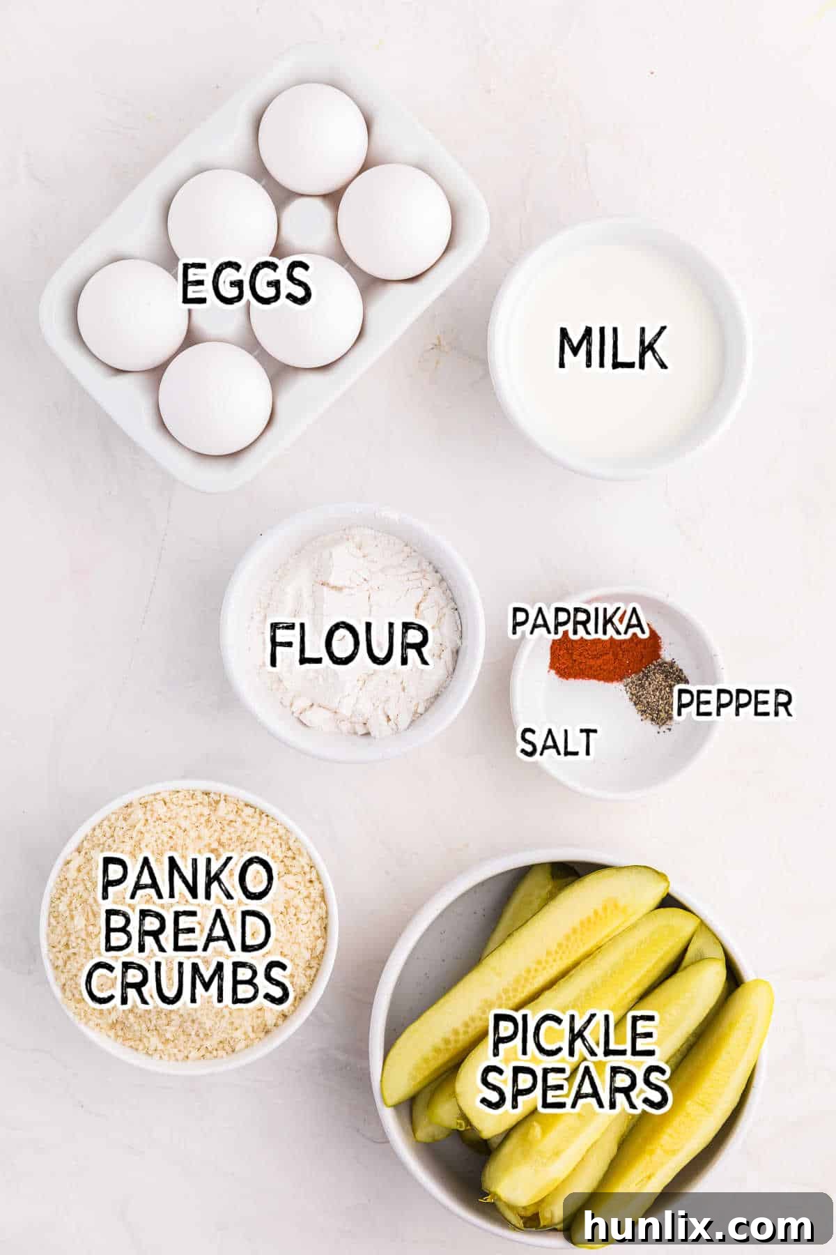 A selection of fresh ingredients including dill pickle spears, flour, eggs, milk, panko breadcrumbs, and spices, laid out on a white counter ready for air fryer fried pickles preparation.