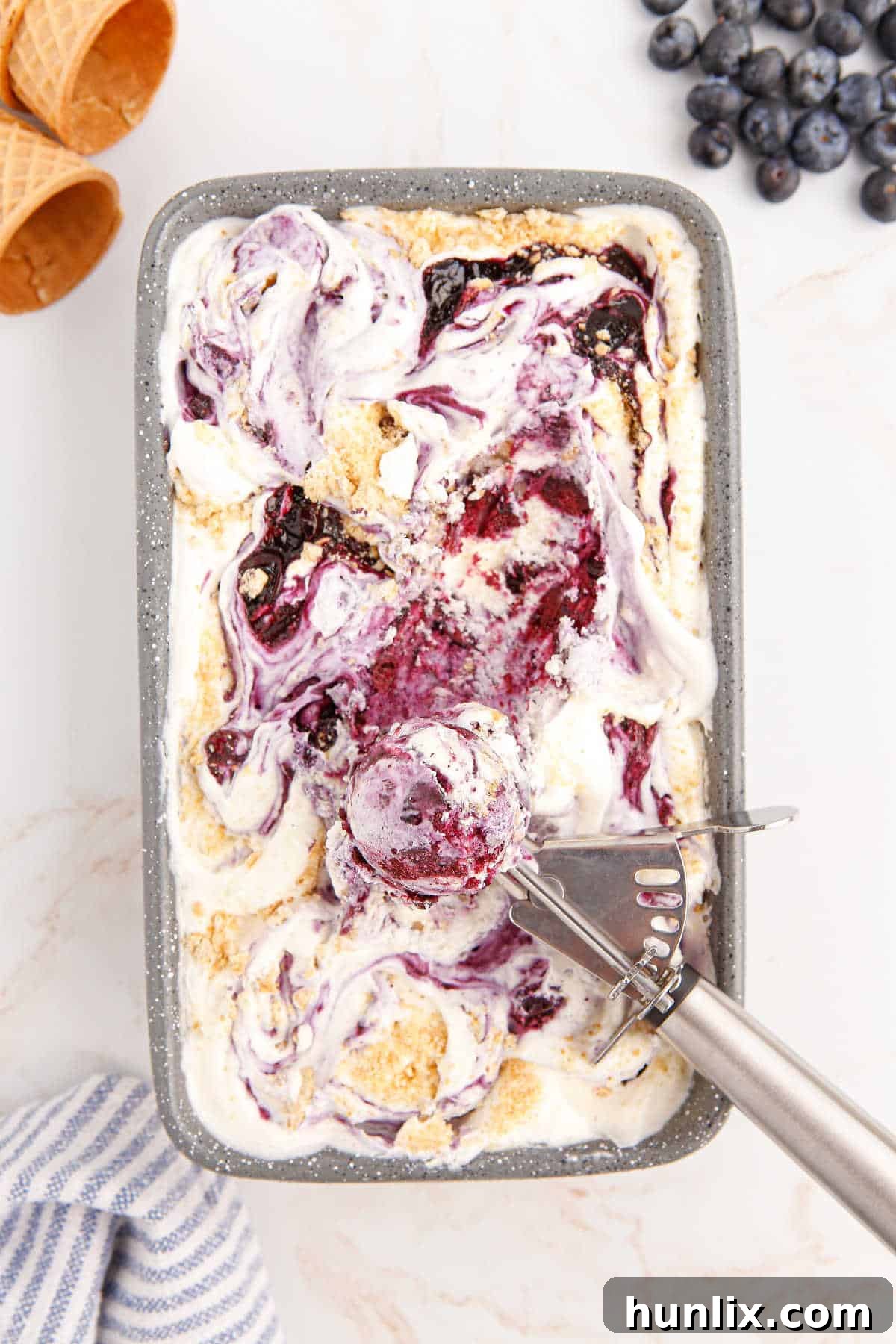 Creamy Blueberry Pie Ice Cream in a loaf pan with an ice cream scoop, showcasing its luscious texture and vibrant blueberry swirls.