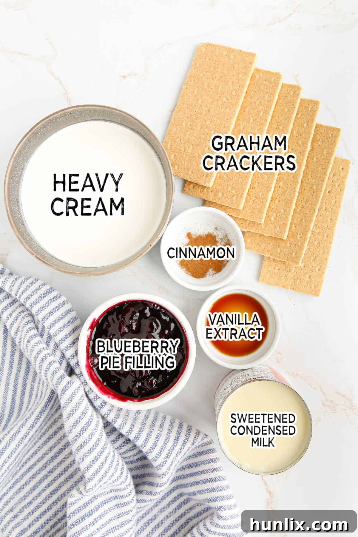 Assortment of fresh ingredients for making homemade blueberry pie ice cream, including heavy cream, sweetened condensed milk, blueberry pie filling, graham crackers, vanilla extract, and cinnamon, neatly arranged on a white surface.