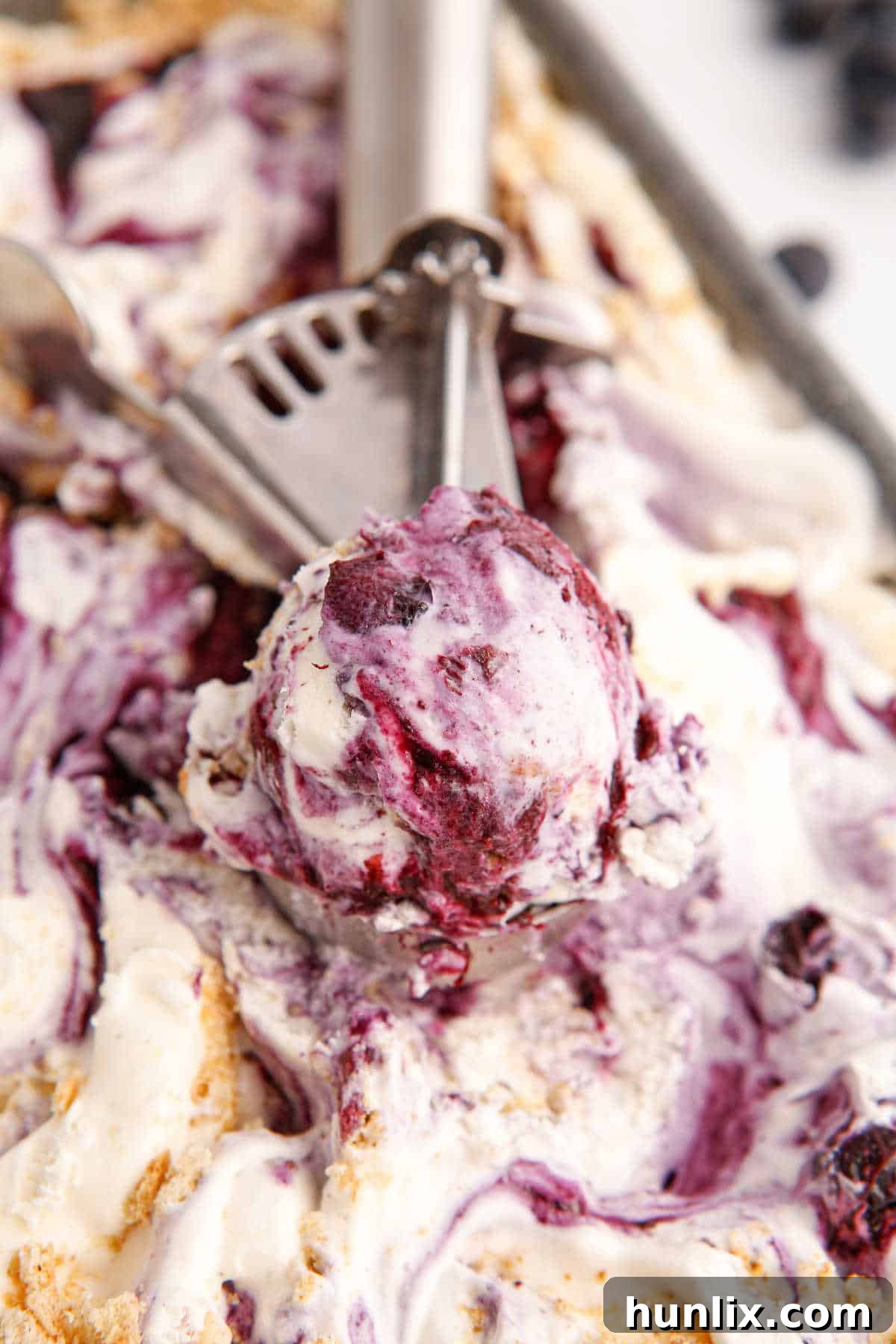 An ice cream scoop digging into a tub of incredibly creamy blueberry pie ice cream, showcasing its rich, luscious texture and the delightful swirls within.