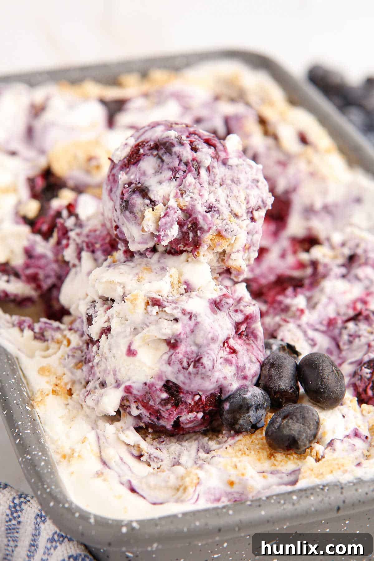 A loaf pan overflowing with creamy blueberry pie ice cream, adorned with several freshly scooped portions and a scattering of bright, fresh blueberries.
