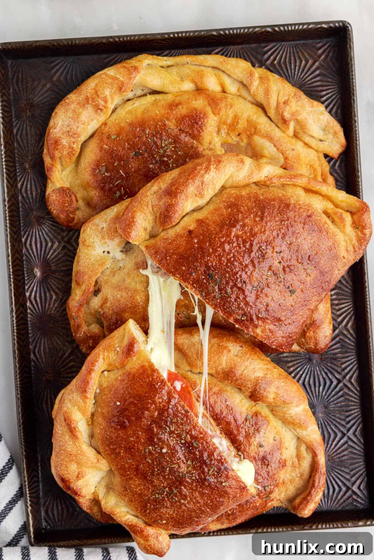 Two meat calzones on a baking sheet, one of which is sliced in half to reveal the rich, melted cheese and meat filling inside.