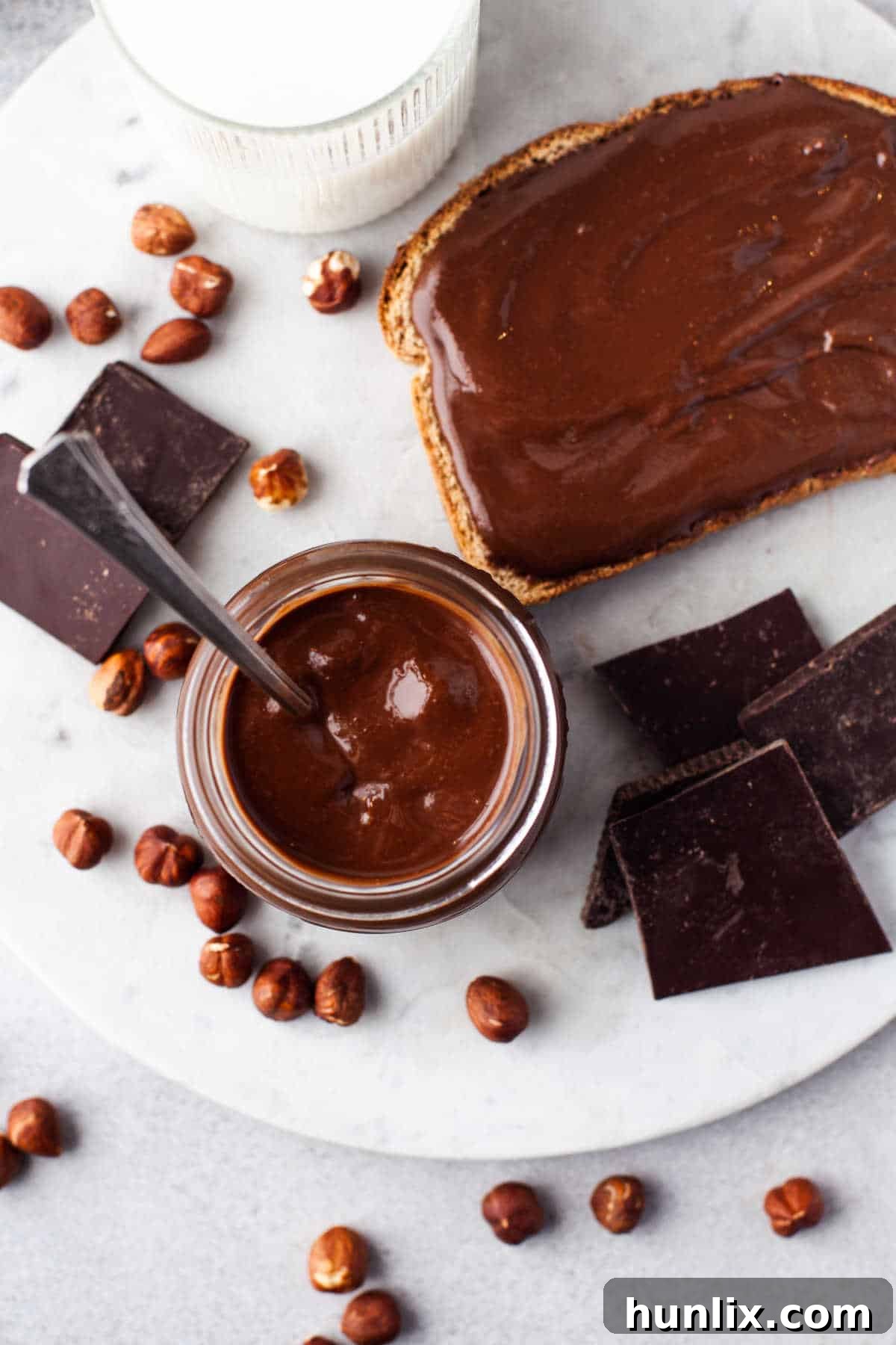 Decadent Homemade Chocolate Hazelnut Spread 7 A close-up of homemade Nutella in a glass jar with a spoon, beside a slice of toast generously spread with the chocolate hazelnut goodness.