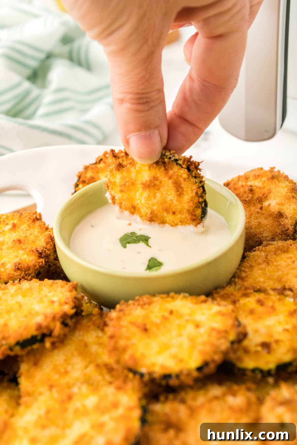 Golden Crispy Air Fryer Zucchini 7 A hand dipping a crispy air fryer zucchini chip into a bowl of creamy white dipping sauce.
