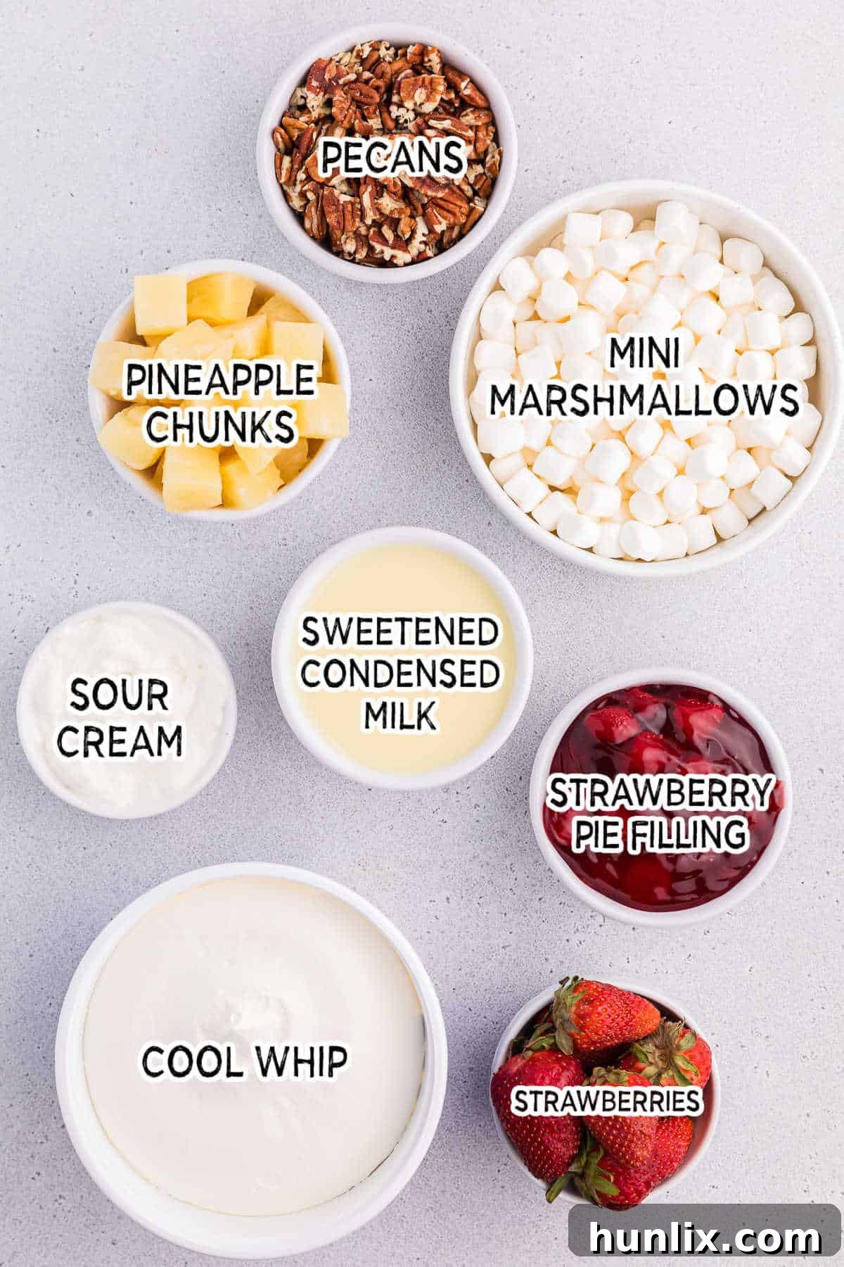 Ingredients laid out on a table to make strawberry fluff salad: strawberry pie filling, sweetened condensed milk, Cool Whip, sour cream, marshmallows, pineapple chunks, pecans, and fresh strawberries.