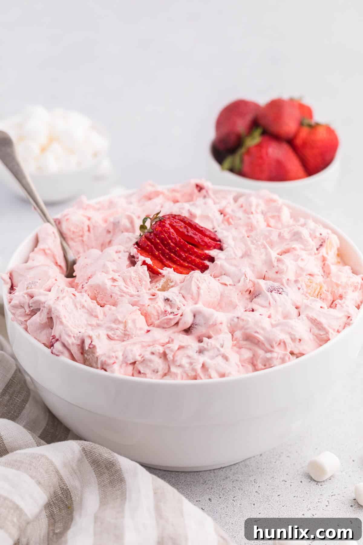 Strawberry fluff salad in a white bowl with a serving spoon, garnished with a few fresh strawberries.