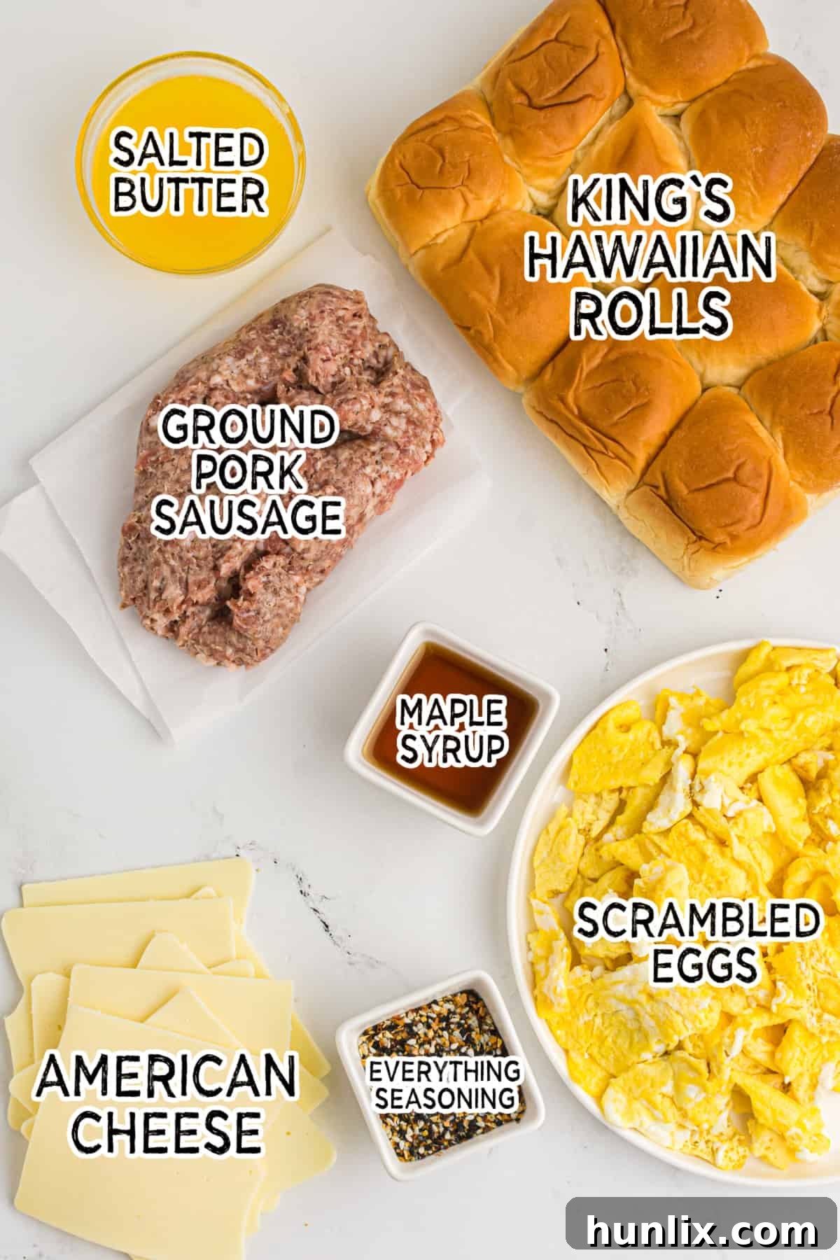 Ingredients for make-ahead breakfast sliders laid out, including Hawaiian rolls, cheese, eggs, sausage, butter, maple syrup, and everything seasoning.