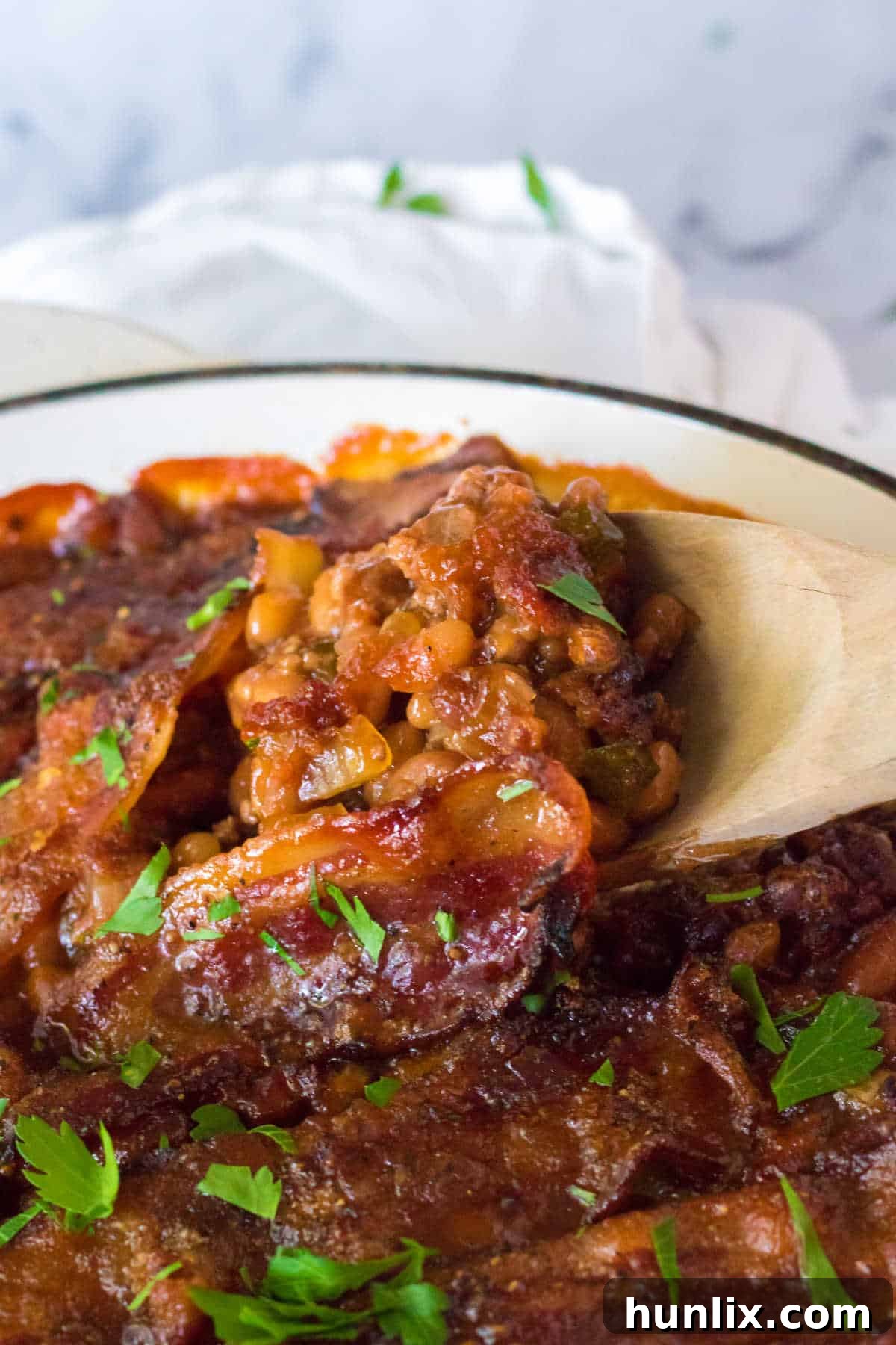 A wooden spoon scooping out baked beans with ground beef from a large pot, showing the rich, thick texture.