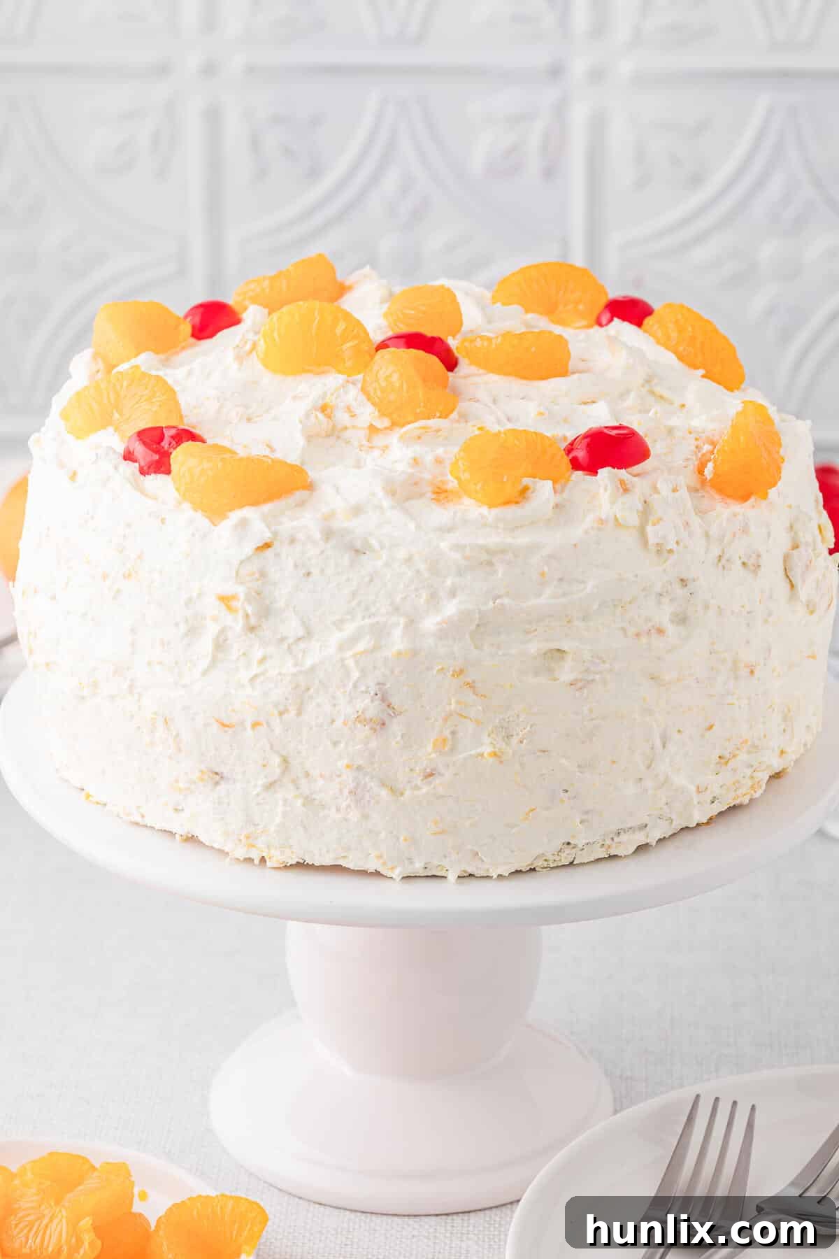 Sunshine Citrus Cloud Cake 2 A vibrant, multi-layered Pig Pickin' Cake on an elegant cake stand, adorned with fresh mandarin orange segments and glistening maraschino cherries. It hints at a delicious, fruity Southern dessert.