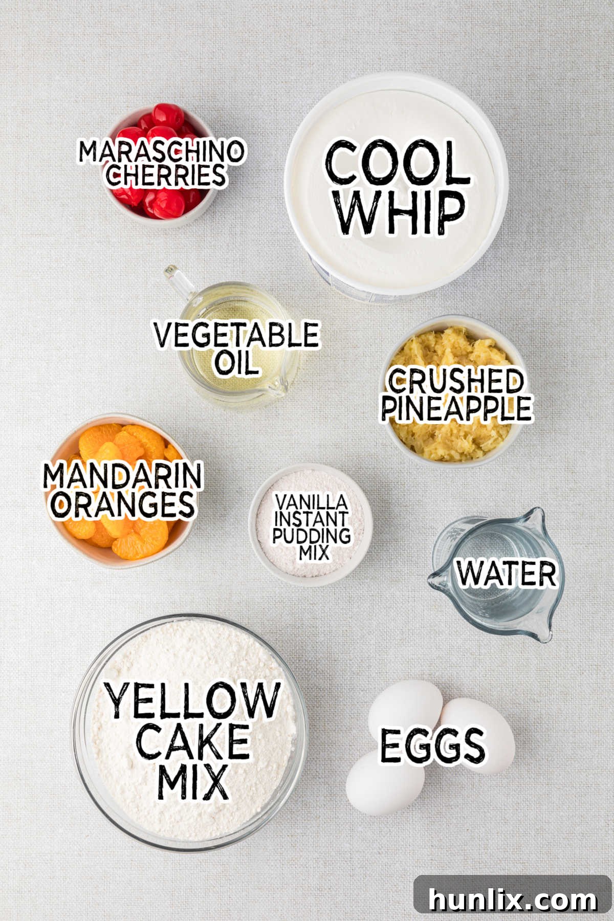 Sunshine Citrus Cloud Cake 3 A flat lay photograph showcasing all the ingredients for Pig Pickin' Cake neatly arranged on a gray background, including a box of yellow cake mix, cans of mandarin oranges and crushed pineapple, eggs, oil, water, pudding mix, and Cool Whip.