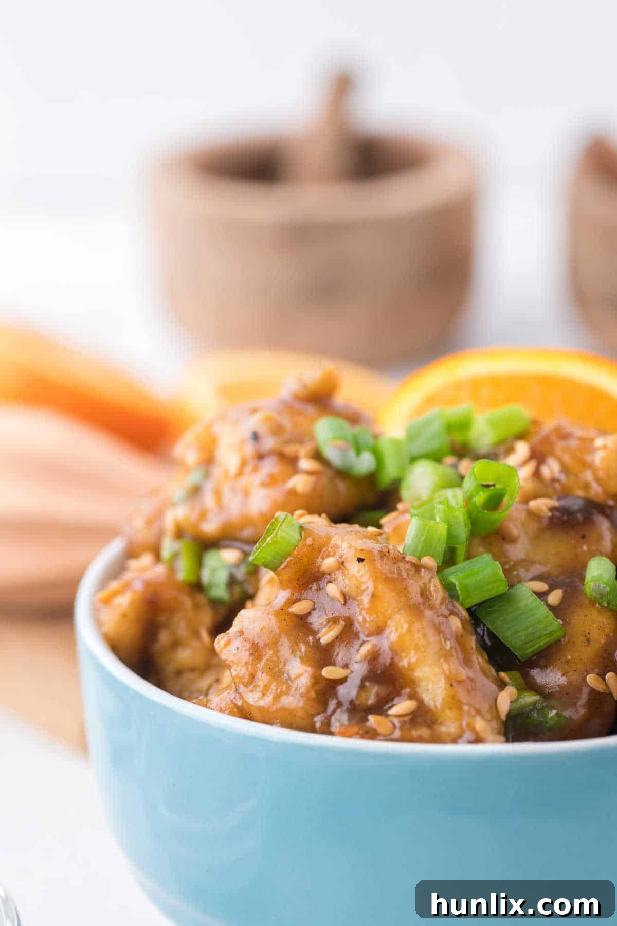 A close-up shot of individual pieces of orange chicken in a blue bowl, highlighting the crispy breading and glossy orange sauce.