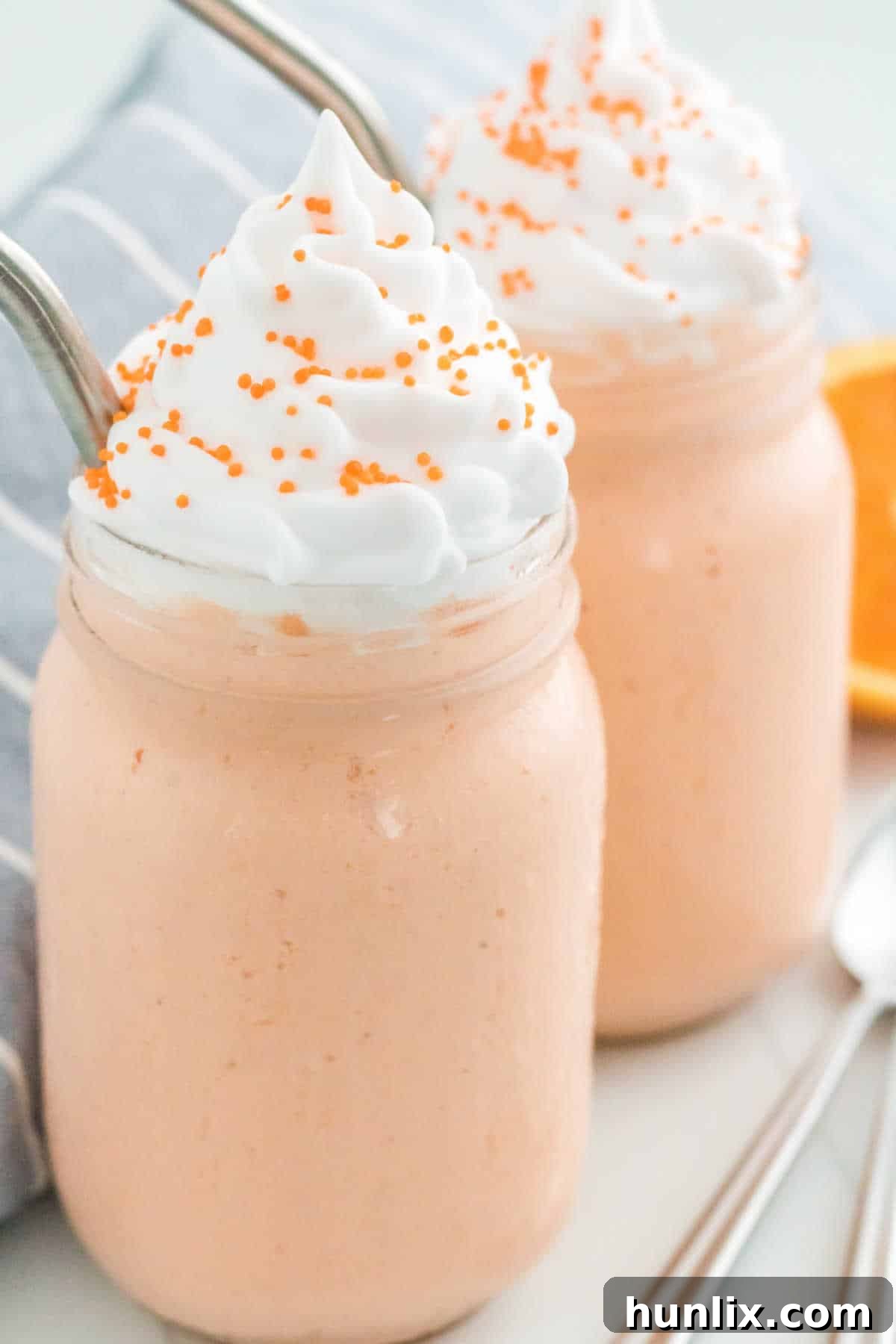 Two boozy orange creamsicle milkshakes with metal straws, topped with whipped cream and sprinkles, presented in tall glasses.