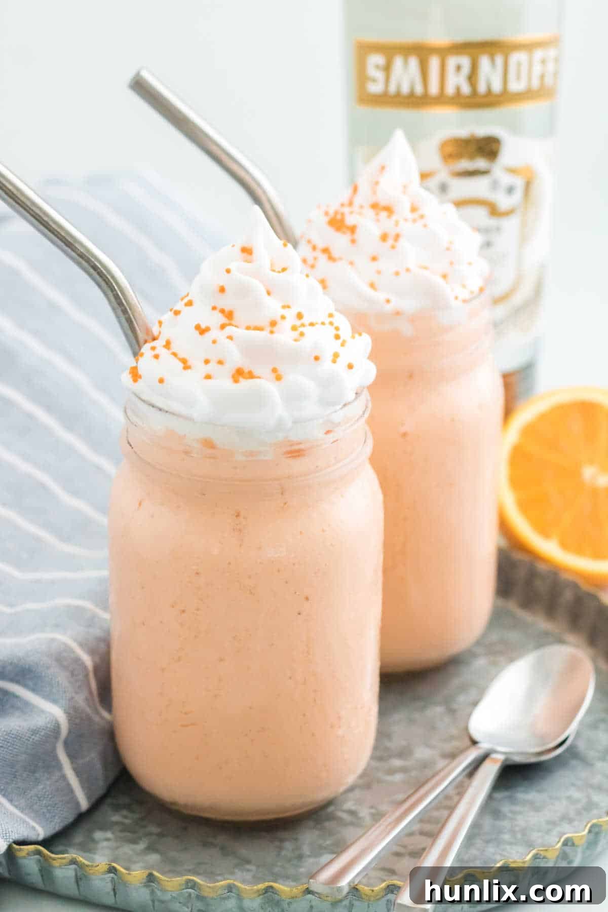 Two boozy orange creamsicle milkshakes garnished with whipped cream and sprinkles, served in tall glasses with metal straws.