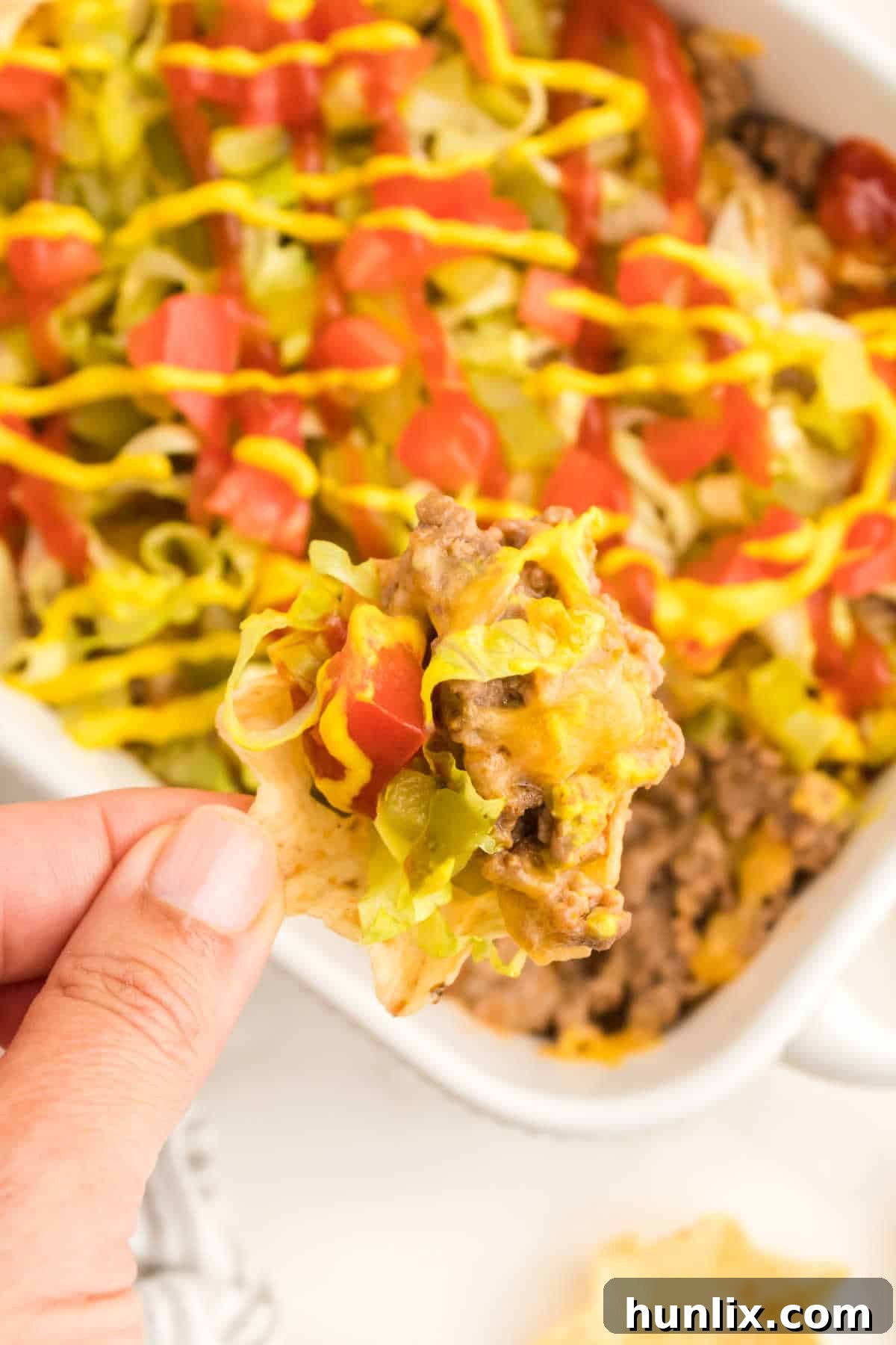 A close-up of a hand holding a crispy tortilla chip adorned with a generous scoop of cheeseburger dip, ready for a delicious bite.