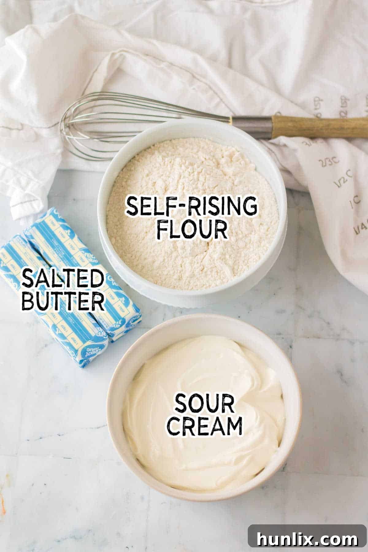 A collection of the three simple ingredients: butter, sour cream, and self-rising flour, laid out on a white marble counter.