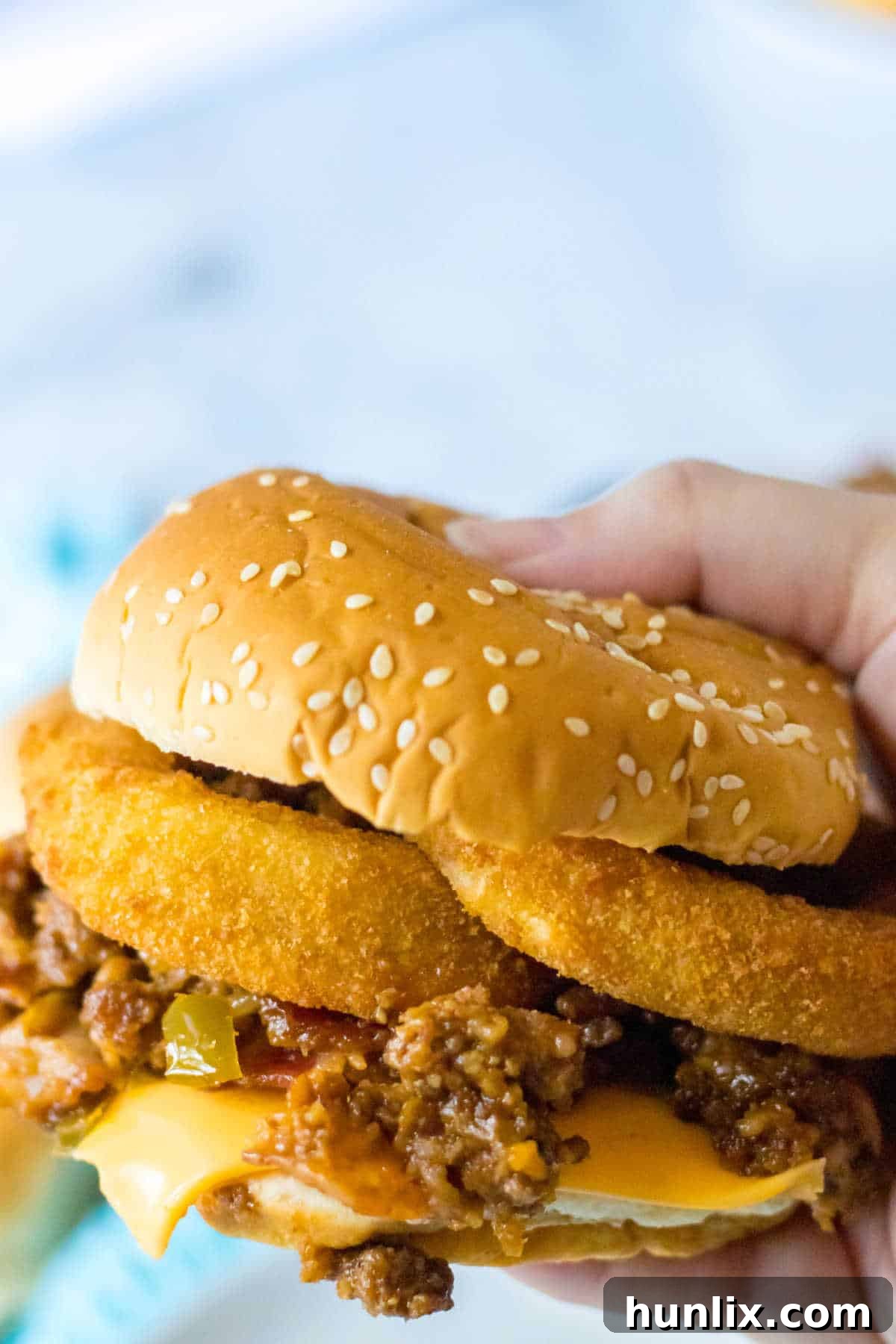 A hand holding a messy, delicious rodeo sloppy joe, highlighting the onion rings and cheese.