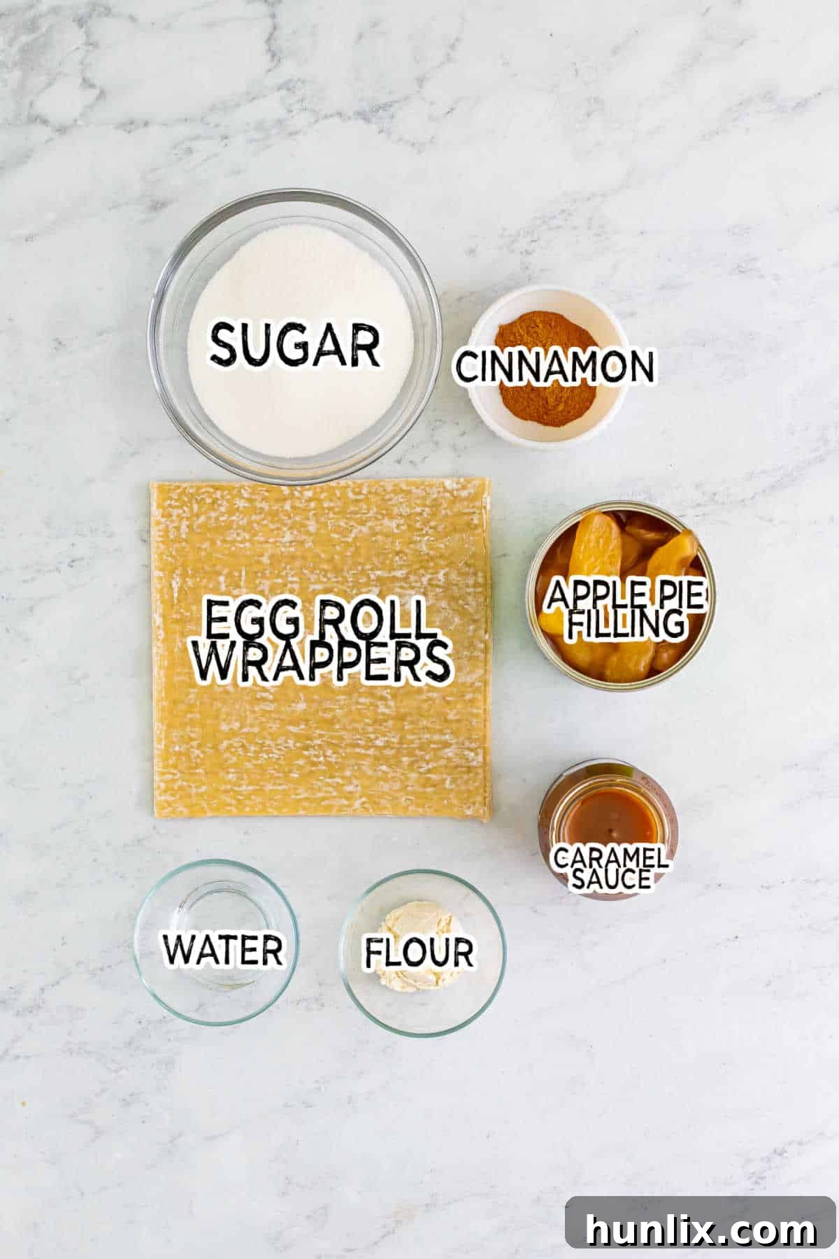 Ingredients for apple pie egg rolls laid out on a marble counter, including egg roll wrappers, canned apple pie filling, sugar, cinnamon, and flour paste.