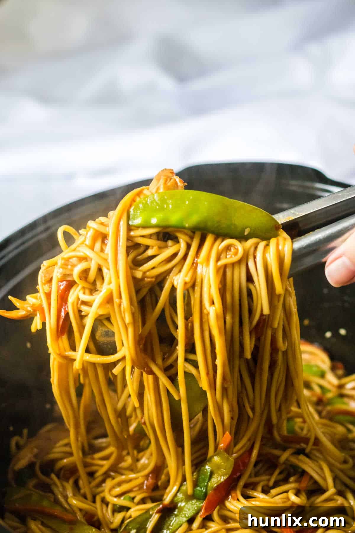 Tongs lifting a generous portion of slow cooker chicken lo mein, showcasing the noodles, chicken, and vegetables.